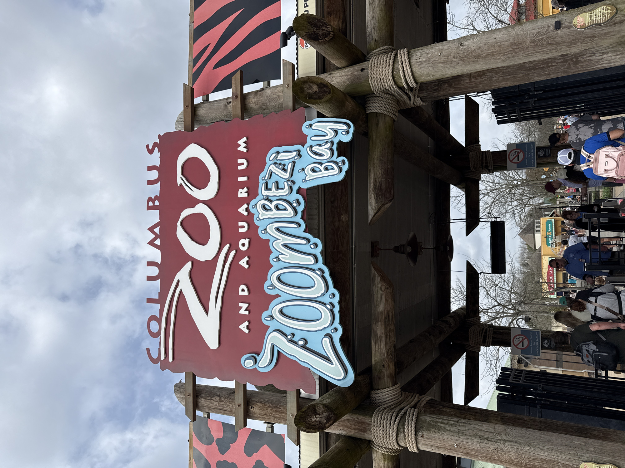 Here's a description of the image:
The image shows the entrance sign to the Columbus Zoo and Aquarium in Powell, Ohio, United States of America. The sign is made of a red background with the words "Columbus Zoo and Aquarium" written in white letters. The words "Zoombezi Bay" are written in blue letters with white accents on a separate sign attached below. The sign is attached to a wooden structure made of logs and rope. In the background, there are people lined up to enter the zoo. There is a sign that reads "Smoke-Free Facility." The sky is overcast.
