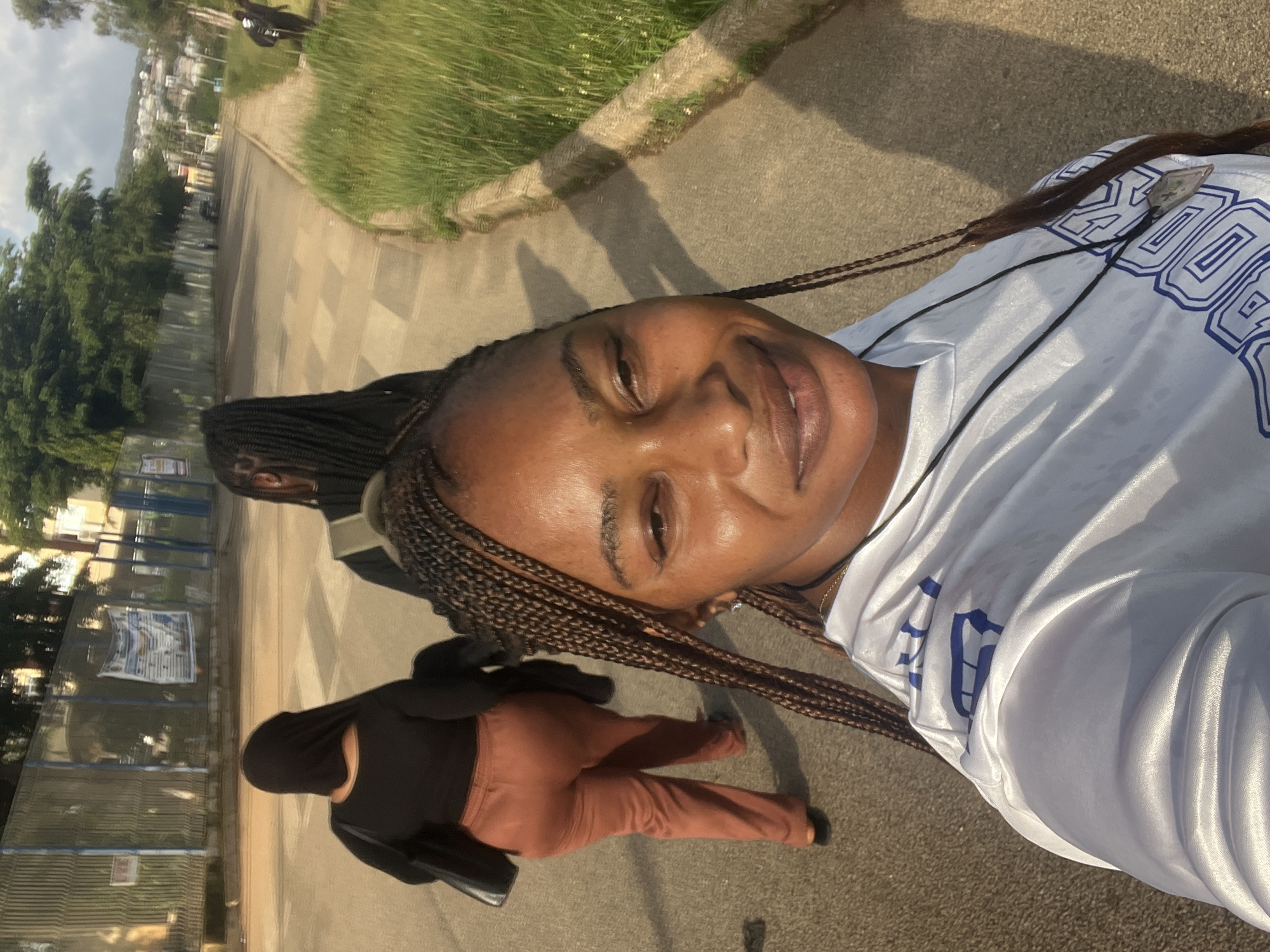 In this eye-level, medium shot taken in Jos, Nigeria, three women are walking on a paved pathway adjacent to a grassy embankment. The woman closest to the camera, in the foreground, is facing the camera and smiling. She has dark braided hair that cascades down her shoulders and is wearing a white t-shirt with blue lettering. To her left, a second woman is walking away from the camera, seen from the back. She is wearing a black hijab and a rust-colored pair of trousers, and she is carrying a black bag over her shoulder. Further in the background, a third woman is also walking away from the camera, her back to the viewer. She has long, dark braided hair and is wearing a dark top and a green backpack. The pathway is a mix of concrete and possibly stone paving, and it is bordered by a tall metal fence on the left. Beyond the fence, trees and buildings are visible. The sky is partly cloudy, and the sun casts shadows on the ground, indicating it is likely late afternoon. The overall setting appears to be a public space, possibly near an educational institution or a recreational area within the city.