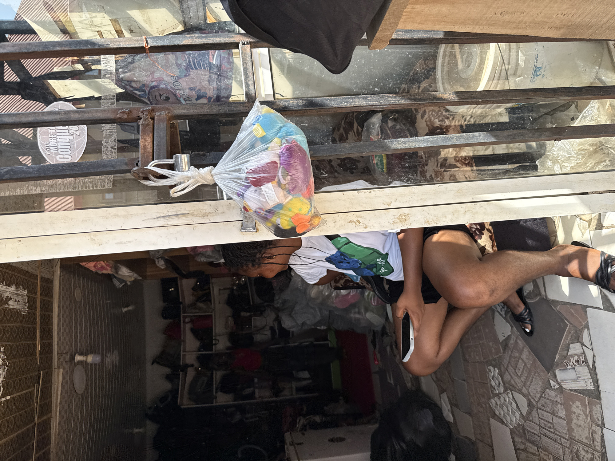 In Abuja, Nigeria, a woman is seated on the ground outside a shop, looking towards her right. She is wearing a white t-shirt with a green and blue design and black shorts. In her hands, she holds a smartphone. A plastic bag filled with colorful toys is tied to a metal gate in front of the shop, with a padlock securing the gate. The shop appears to be a retail establishment, with shelves of bags and other merchandise visible inside. The ground is paved with tiles, some of which are broken. The scene suggests a typical street-side business environment in the city.