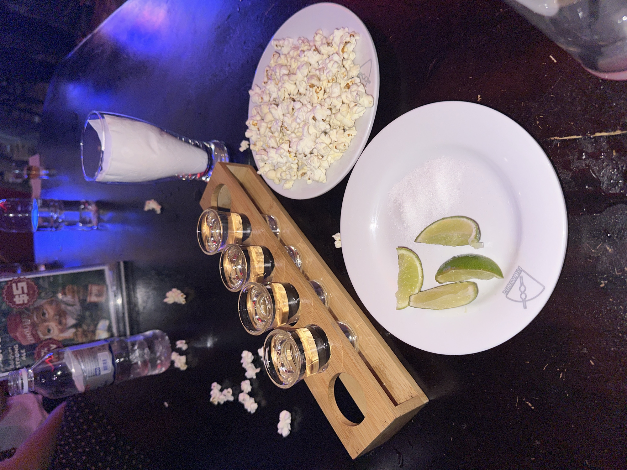 The image captures a dark, reflective tabletop, likely in a bar or restaurant, with blue and purple ambient lighting creating a moody atmosphere. In the foreground, a white plate holds several lime wedges and a pile of white salt. To the left, a wooden tray supports four shot glasses filled with a clear liquid, presumably liquor. Next to the shot glasses, a plate is piled high with popcorn. Further back, a tall glass holds a white paper napkin, and a water bottle stands near a sign with an image of a gnome. Scattered across the table are small pieces of popcorn. The overall scene suggests a casual gathering with drinks and snacks. There is no indication of the specific location within Khan Chamkar Mon, Cambodia.