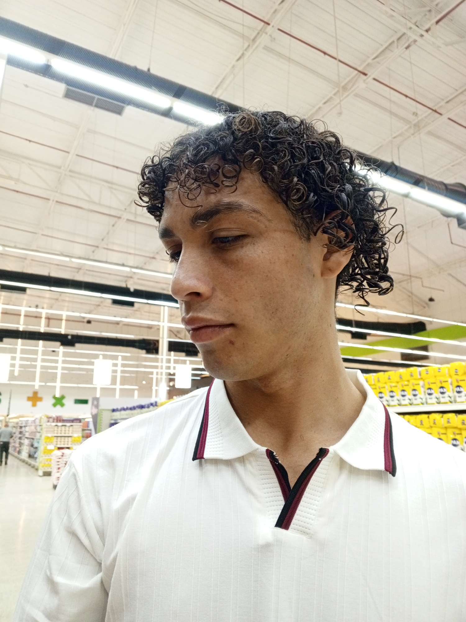 A medium shot shows a person with curly hair and a white polo shirt with red and black trim, looking downward in a brightly lit supermarket. Rows of shelves with goods are visible in the background, typical of a retail shopping environment. The setting suggests an indoor space, possibly located in Porlamar, Venezuela, given the prompt's information. The image focuses on the individual's appearance and general surroundings, without highlighting specific actions or interactions.