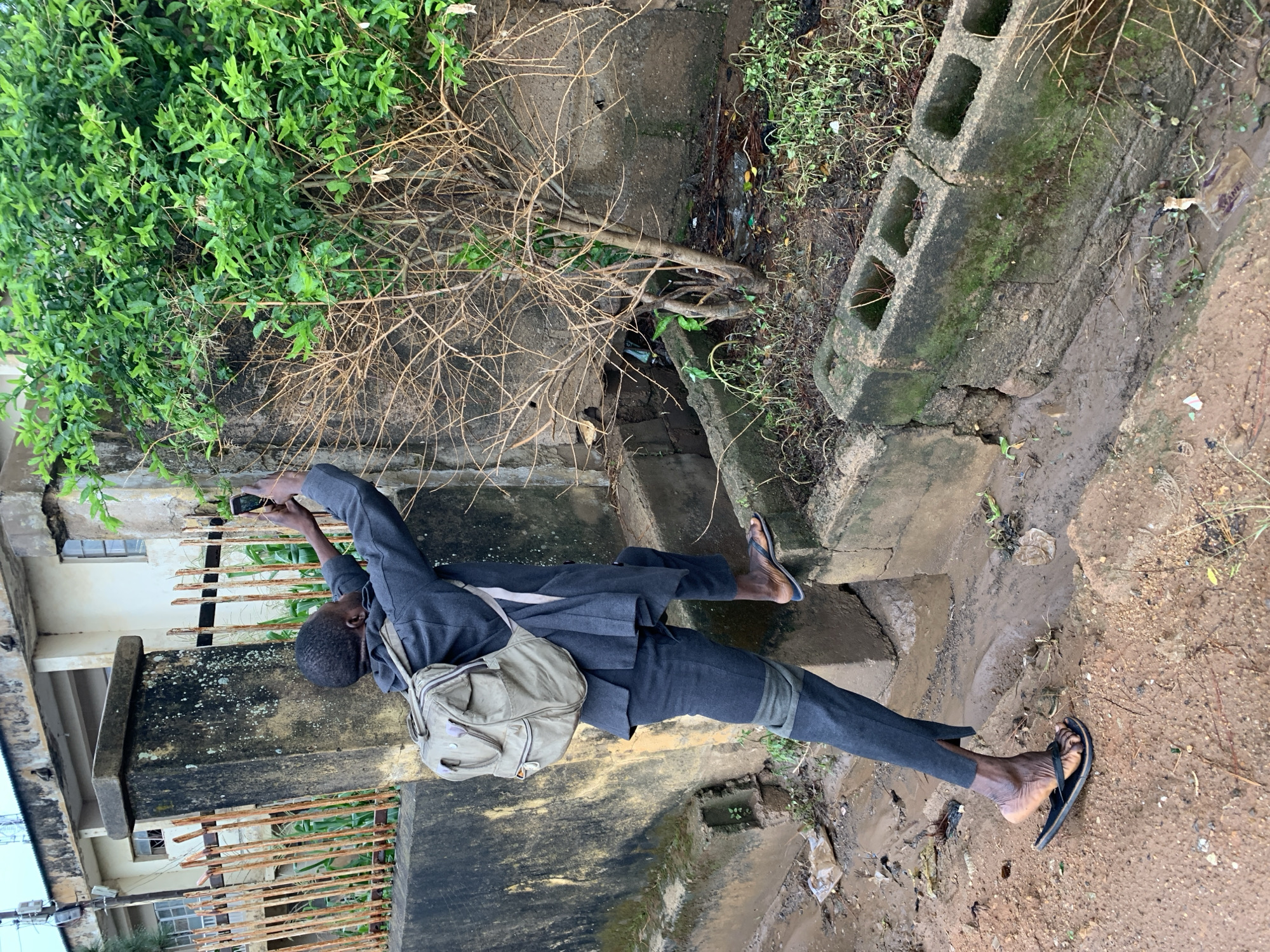 This media file captures an outdoor scene in Rafin Par, Nigeria, featuring a person interacting with their surroundings. The primary subject is an individual, likely male, with dark skin and short hair, positioned slightly off-center, facing towards the left. They are dressed in a dark grey long-sleeved shirt or jacket, dark grey trousers, and black flip-flops, with a light-colored backpack or shoulder bag slung over their left shoulder.
The person is actively holding a dark-colored smartphone with both hands, extended upwards, appearing to be taking a picture or video of the area in front of them, or reviewing content on the device.
The setting is characterized by uneven, muddy ground, indicative of damp conditions or recent rain. Lush green foliage dominates the upper left and parts of the background, contrasted by a mass of dry, thorny branches extending across the mid-ground. Concrete structures, including a broken concrete block with moss growth and what seems to be a drainage ditch or culvert, are visible. In the background, a building with windows and rusted metal bars or a fence can be seen, suggesting an urban or semi-urban environment. The lighting suggests it is daytime, possibly overcast, as there are no harsh shadows. A small white wrapper with the text "WANE" is discernible on the muddy ground near a concrete block.