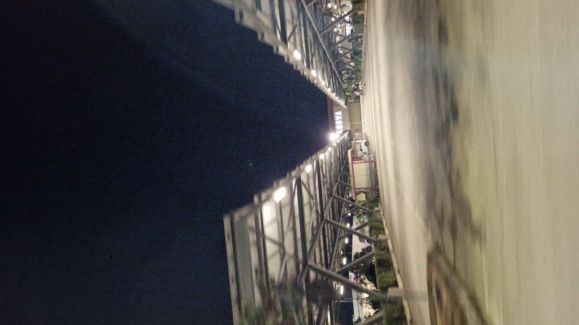 The image depicts a nighttime scene in a parking lot covered by solar panel structures. The structures, characterized by their geometric metal framework, are illuminated by overhead lights. In the background, a building with the word "ANOVA" displayed prominently on its facade is visible. A few people are walking in the distance, and a white car is parked near the building. The parking lot is paved, and there are rows of shopping carts visible under one of the solar panel structures. The sky is dark, suggesting it is late at night. The blur in the foreground indicates movement, possibly from the camera or the vehicle from which the image was taken. This scene is located in the city of Taranto, Italy.
