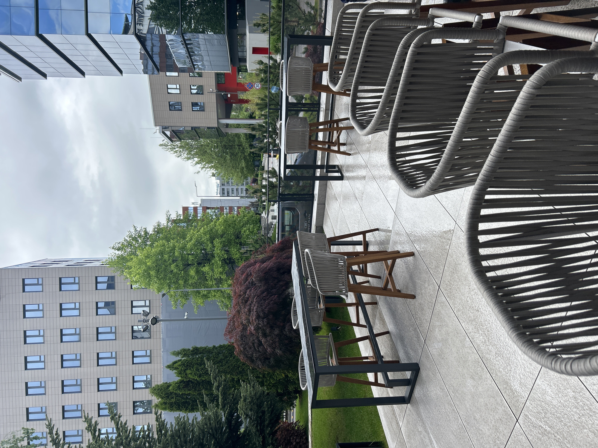 The image shows an outdoor seating area, likely a terrace or patio, located in an urban setting. The furniture consists of several high tables and matching chairs with woven seats and wooden legs. The tables have gray frames and reflective surfaces. The area is paved with light-colored, textured tiles.
In the background, there are multiple buildings of varying heights and architectural styles. Some buildings feature glass facades, while others have more traditional window designs. There are also trees and other greenery visible, suggesting a park or landscaped area nearby. The sky is overcast, indicating a cloudy day.
Based on the signs and building styles in the background, the location is likely Sofia, Bulgaria. The "30" speed limit sign is a standard European road sign. The overall appearance of the buildings and the urban planning align with the characteristics of Sofia.