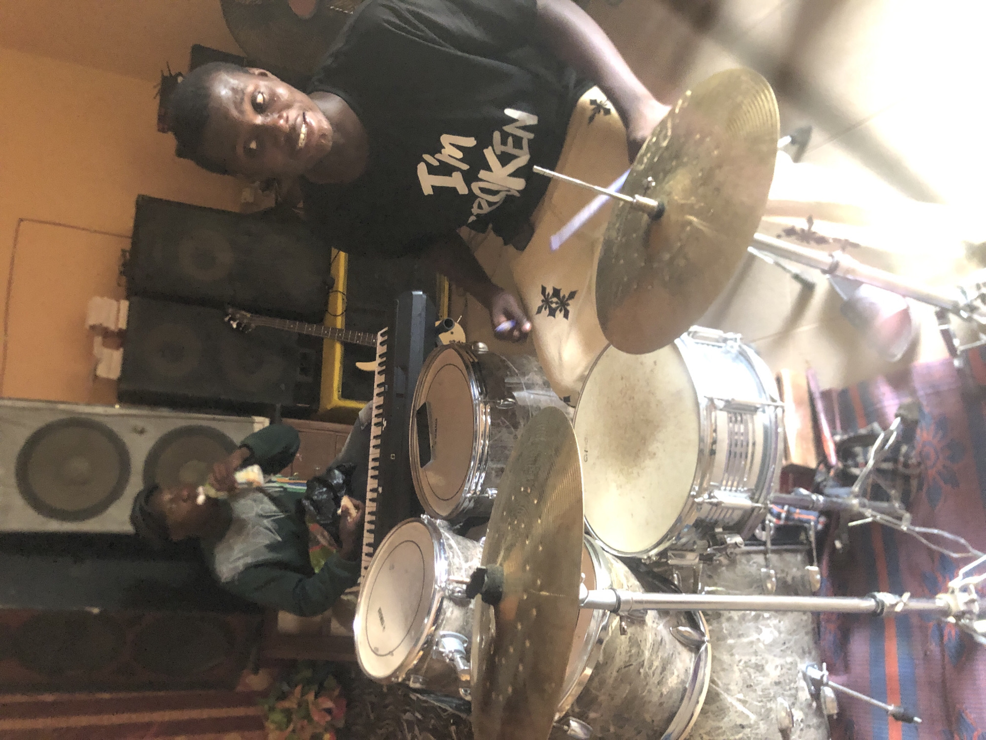 The image shows a man playing a drum set in what appears to be a music studio or practice room in Nkwelle Ezunaka, Nigeria. The man is wearing a black t-shirt with the words "I'm Broken" printed on it. He is holding drumsticks and appears to be striking a cymbal. The drum set consists of several drums and cymbals, all in a silver finish. In the background, there are speakers, a guitar, and a keyboard. Another person is sitting behind the speakers, possibly eating. The walls are painted in a muted orange hue. A striped blanket is draped on the floor. The environment is cluttered and suggests a lived-in space where music is frequently played.