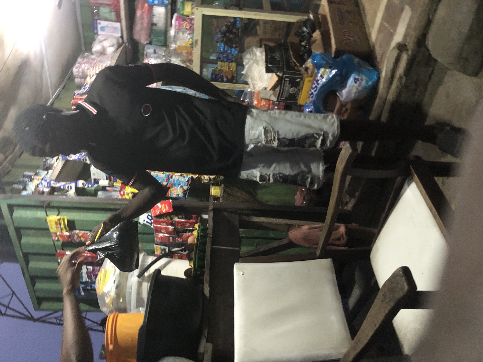 In Ado, Nigeria, at dusk, a man in a black polo shirt and ripped denim shorts stands in front of a small store. He is receiving a black plastic bag from another person whose arms are visible on the left side of the frame. The store is stocked with various items, including packaged goods on shelves and in baskets. A white-cushioned chair is positioned to the left of the man. The environment suggests a busy marketplace or a roadside shop.