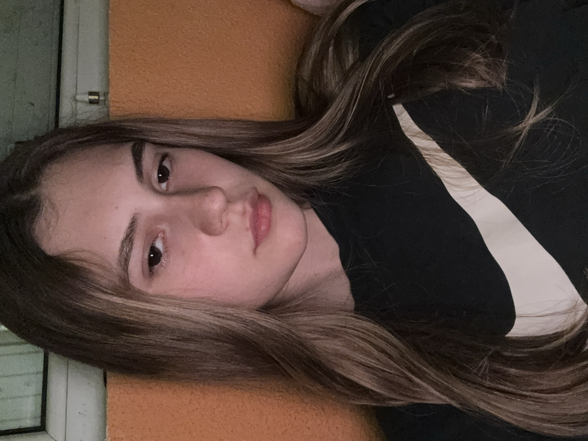 A young woman is depicted lying down, resting her head on an orange, textured surface. Her long, wavy hair, dark brown with noticeable lighter highlights, is spread around her. She has dark brown eyes and a neutral expression, with her lips slightly parted. She is wearing a black t-shirt with a prominent white Nike swoosh logo visible on the right side of the frame, over her chest or shoulder area.
The background suggests an indoor setting. To the left of her head, there's a greenish-grey vertical surface, next to a white pipe or frame that has a small metallic clip attached. Below the orange surface, on the far left, a dark window frame is partially visible. The lighting is soft and even, indicating an indoor environment. The woman's pose and expression convey a relaxed and calm mood. The only visible text is the Nike logo. The scene takes place in Panagyurishte, Bulgaria.