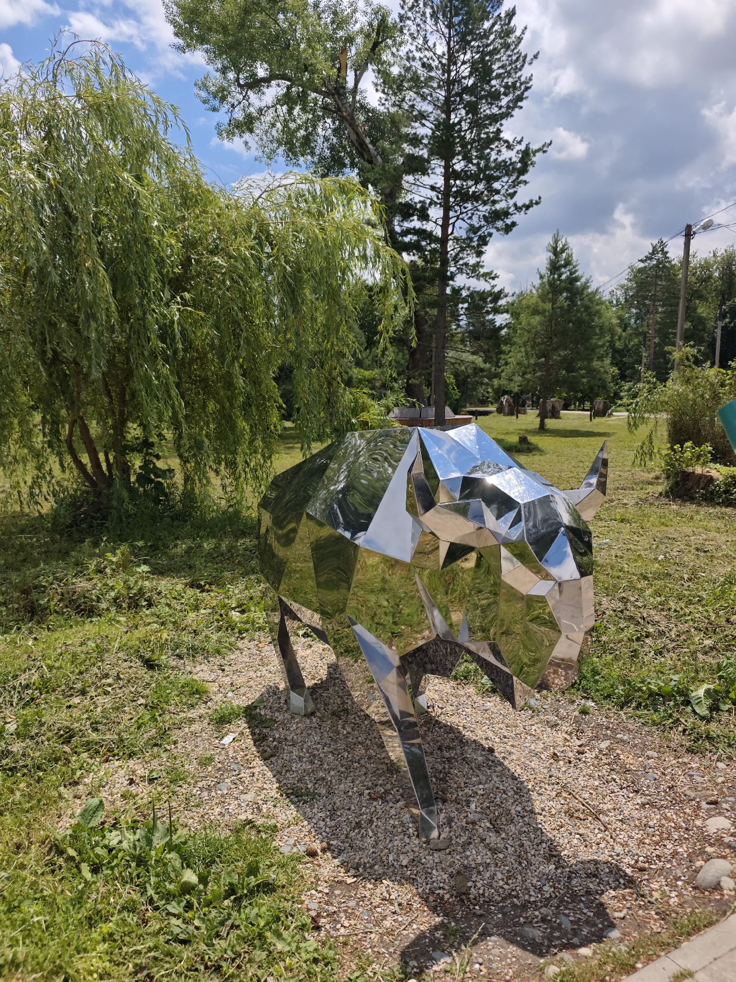 An outdoor scene in Cherkessk, Russia, features a polished, geometric metal sculpture of a bison. The sculpture is positioned on a gravel path, casting a distinct shadow on the ground. Lush green foliage, including a weeping willow tree on the left, surrounds the path and sculpture. In the background, various trees, including pine trees, dot the landscape under a partly cloudy sky. The image captures a serene park-like setting with natural elements and a striking modern art piece.