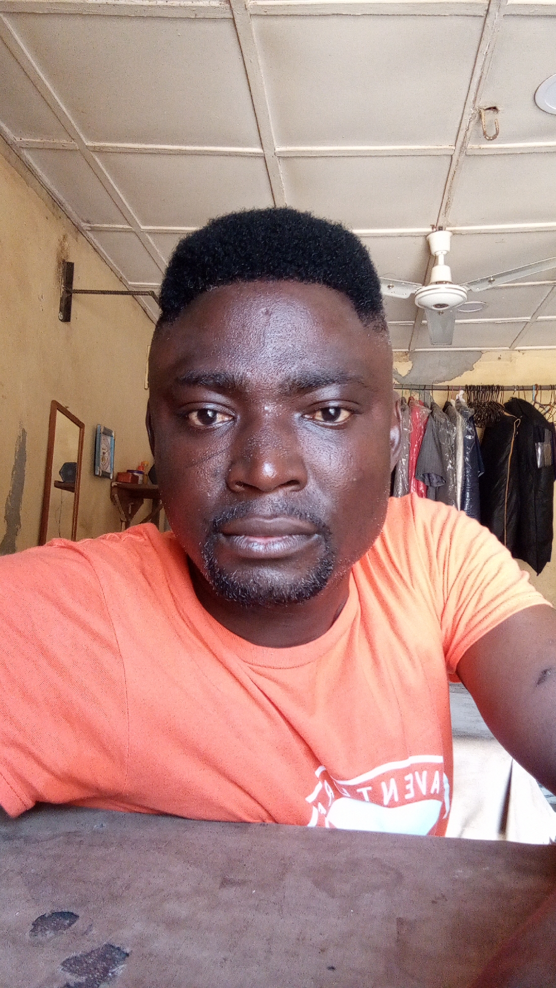 A close-up shot depicts a dark-skinned man with short black hair and a trimmed beard, wearing an orange t-shirt. The setting appears to be indoors, with a white ceiling adorned with light fixtures and a ceiling fan. In the background, clothing hangs on a rack. The scene is set in Sabon Tasha, Nigeria.