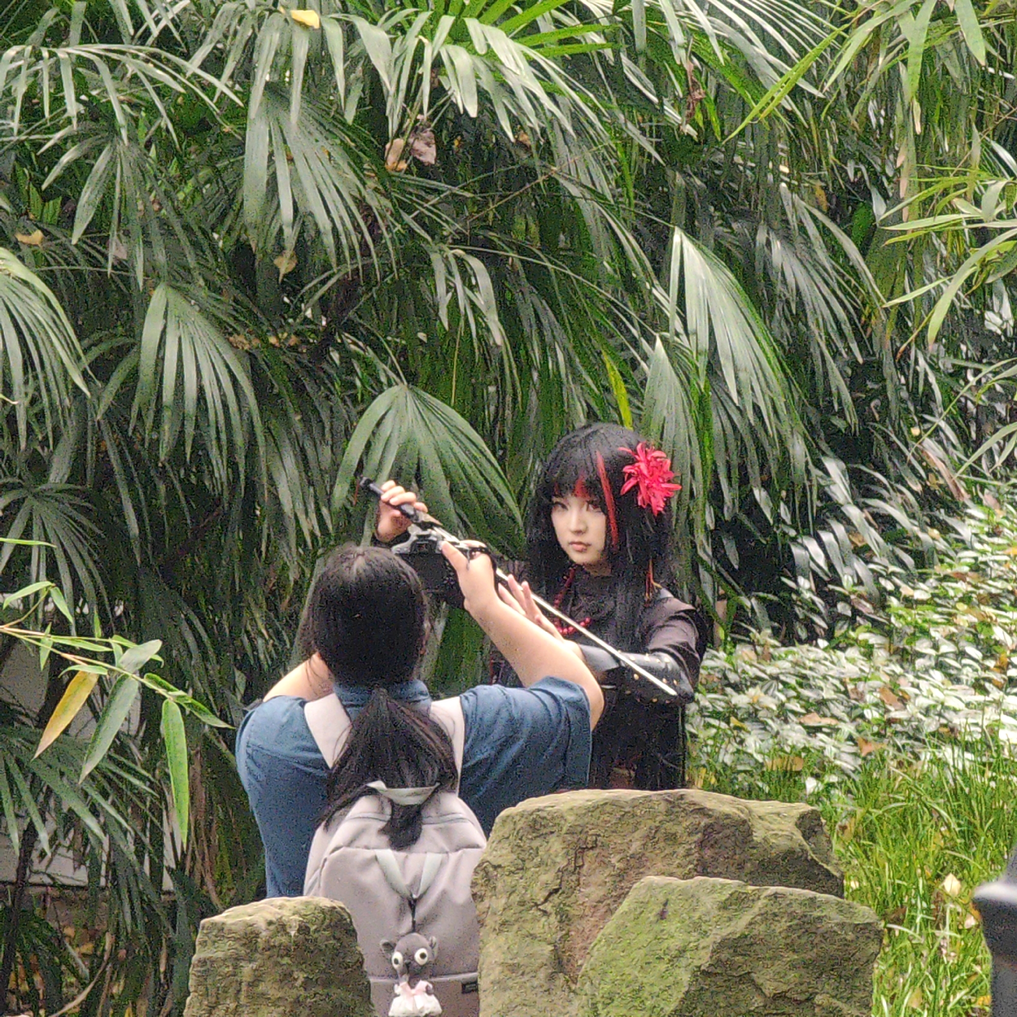 In Yuzhong District, China, a person with a grey backpack stands facing a person dressed in black clothing and sporting a large red flower in their dark hair. The person in black holds a sheathed sword. Green foliage, including bamboo and palm-like leaves, forms the backdrop, with moss-covered rocks in the foreground. The person with the backpack appears to be adjusting the sword or interacting with the person holding it. A small stuffed animal is attached to the backpack.