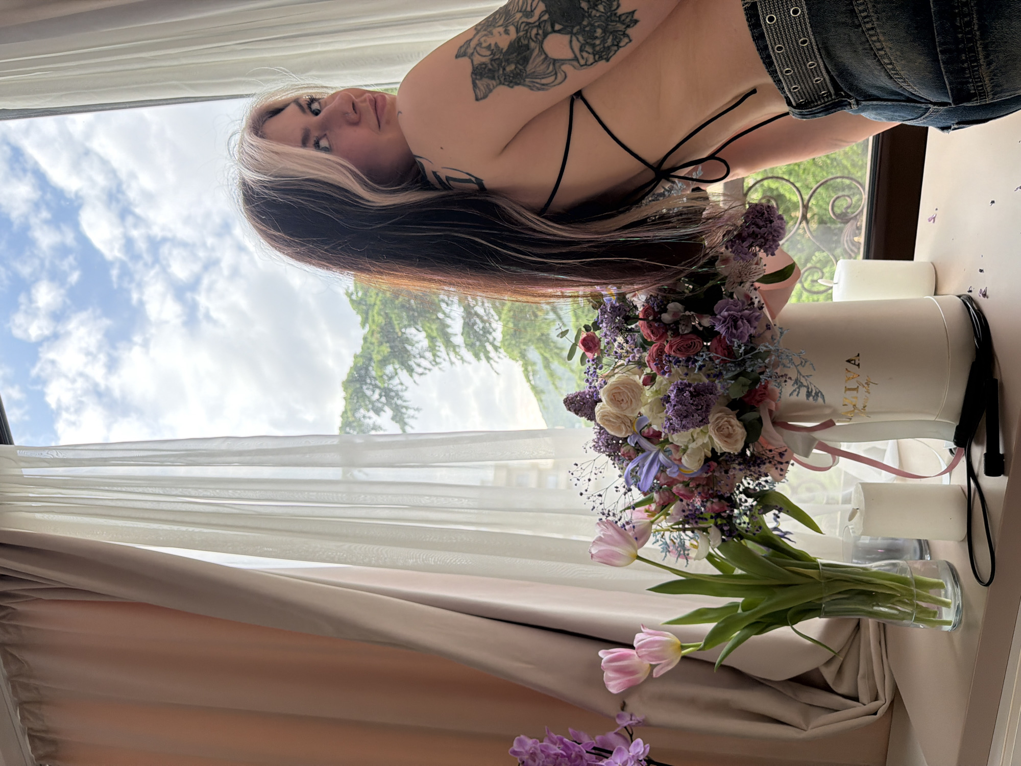 A portrait-style photograph captures a woman posing indoors by a large window, surrounded by floral arrangements. The woman, with long dark hair featuring blonde highlights, is turned away from the camera, looking over her right shoulder with a neutral expression. She wears a black backless halter top and blue denim jeans or shorts with silver eyelet detailing on the waistband. Her right arm and shoulder are adorned with intricate black tattoos depicting floral and figurative designs.
To her right, a large, elaborate bouquet of various flowers including pink, white, and red roses, purple blossoms, and blue bell-shaped flowers is arranged in a white hat box. Gold text, partially visible as "FLORALIVA", is printed on the side of the box. To her left, two light pink tulips stand in a clear glass vase, with another cluster of purple flowers visible at the bottom. Two white pillar candles are placed on the surface next to the hat box. A black cable or strap lies near the candles.
The setting appears to be a brightly lit room with natural light coming from the large window behind her. Sheer white and light brown curtains frame the window. Through the window, a cloudy sky and lush green trees or foliage are visible, suggesting a scenic outdoor view, possibly indicative of the Budva, Montenegro location context. The light suggests it is daytime.