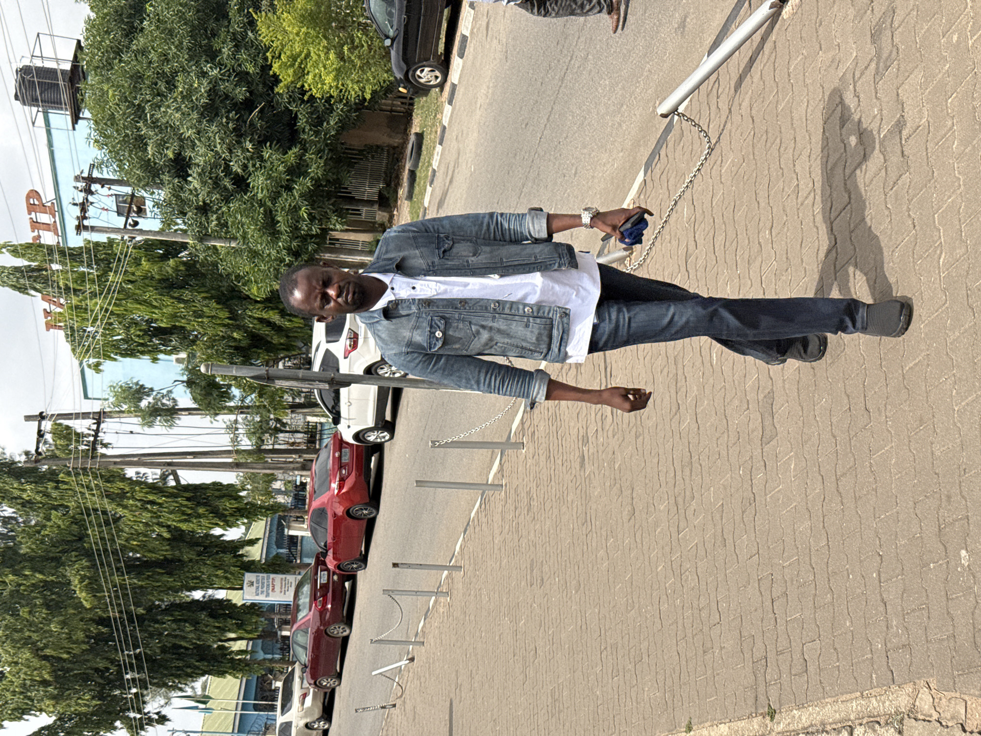 A man stands on a brick paved sidewalk next to a street lined with parked cars. He is wearing a denim jacket, white shirt, and dark pants. He holds a dark-colored object in his right hand. To his left, a row of silver posts connected by chains runs along the sidewalk, separating it from the street. In the background are trees, parked cars (red and white), a building with the letters "IIP" visible, and utility poles. The location is possibly Abuja, Nigeria. The sky is overcast.