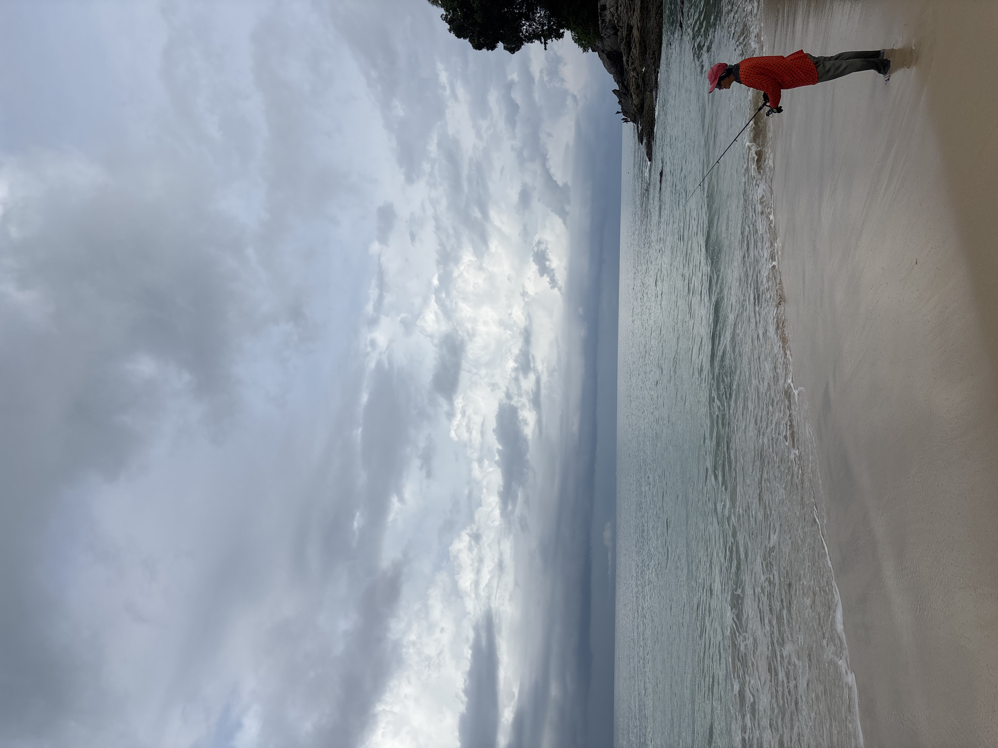 A coastal scene in Choeng Thale, Thailand shows a person fishing at the edge of the water. The person, wearing an orange jacket and pink head covering, is holding a fishing rod, which extends into the water. The sky is overcast with gray clouds. A sandy beach is to the right, with the water to the left. A cliff or rocky outcropping with greenery is situated in the upper-right area of the frame. The tide is receding onto the shoreline, causing small white-watered waves.