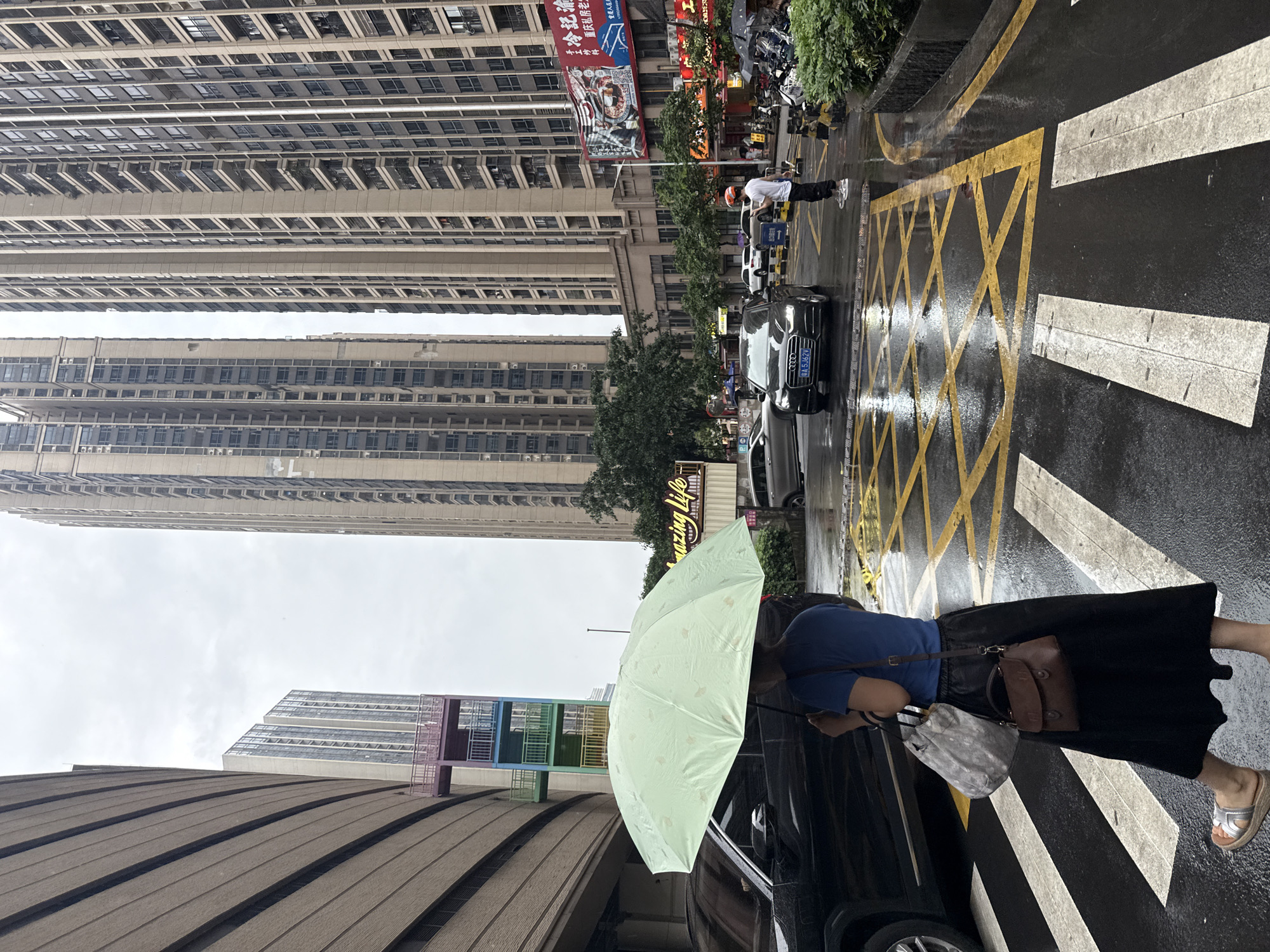 A light green umbrella with a subtle pattern is held by a woman from the back, obscuring her face. She is wearing a blue t-shirt and a long black skirt, with silver sandals on her feet. She carries two bags: a larger, light gray patterned bag over her left shoulder and a smaller, dark brown leather satchel in her right hand. The woman is standing on a wet crosswalk, with yellow "box junction" markings on the asphalt behind her. A black car is visible to her left, and a dark SUV is partially visible to her right. In the background, two tall, modern residential buildings dominate the skyline. To the left of these buildings, a distinct structure with colorful external staircases (green, blue, pink) is visible. The street is wet and reflective, suggesting recent rainfall. A white car is stopped in the road ahead of the woman, and a man in a white shirt and black pants, wearing an orange helmet, is standing on the right side of the road, also looking towards the traffic. The scene appears to be a street in Haizhu District, China, on an overcast day.