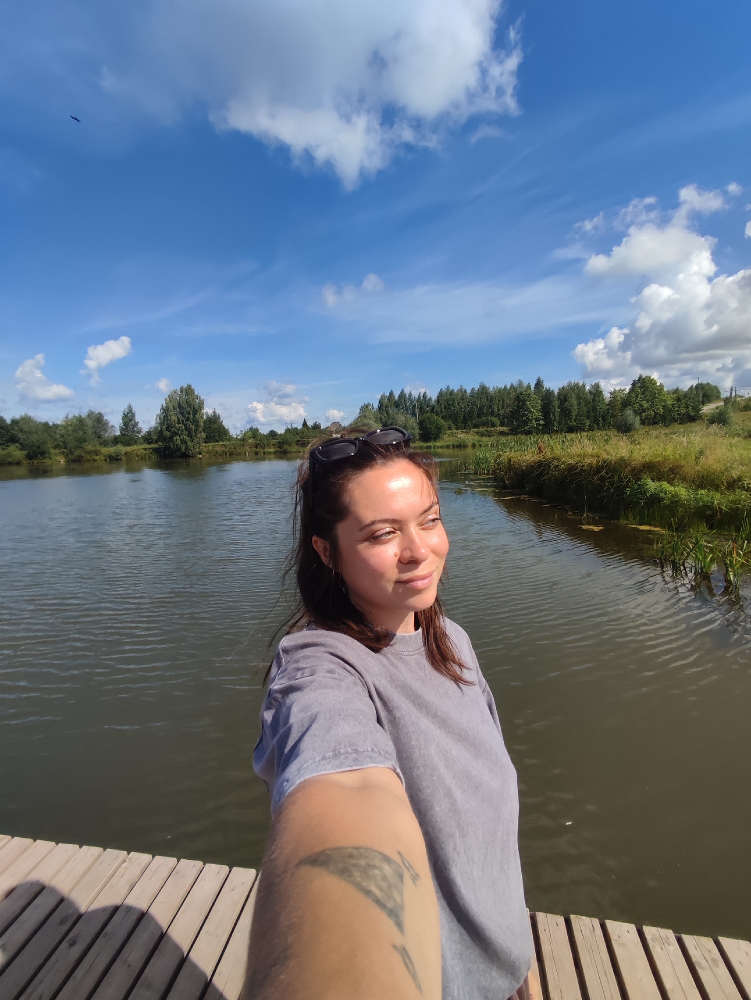 A woman is taking a selfie on a wooden pier next to a body of water. She has dark hair and is wearing a grey t-shirt and sunglasses on her head. In the background, there is a body of water, trees, and a blue sky with white clouds. The scene appears to be in a natural, outdoor setting, likely in the city of Foky, Russia, given the context. The image is taken from a low angle, with the woman's arm extended to capture the selfie. A bird is visible flying in the upper left portion of the sky.