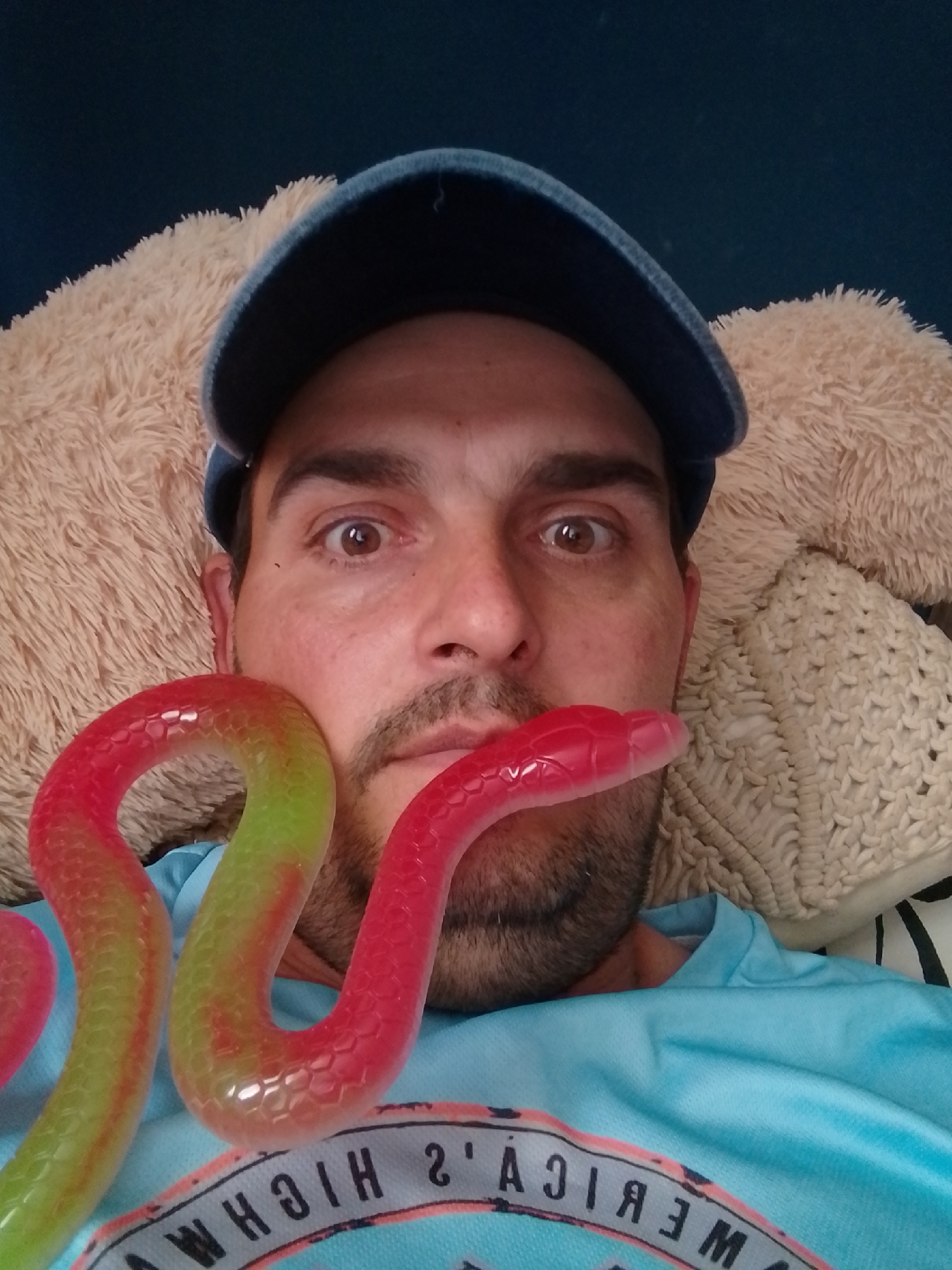 A man wearing a blue baseball cap and a blue t-shirt is pictured from the chest up. He has a mustache and stubble. A colorful, segmented, snake-shaped toy is placed in front of his face, with one end near his mouth and the other curving around his neck. The background consists of soft, textured fabrics in beige and cream colors. The image appears to be a selfie taken indoors. There is no information to suggest this image is located in Wedel, Germany.