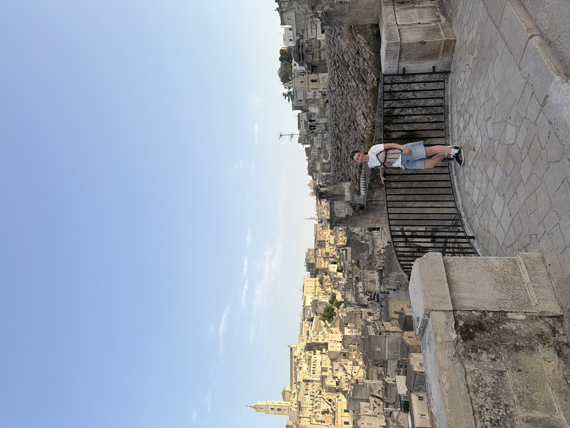 A young woman stands on a stone-paved overlook in Matera, Italy, with the ancient cityscape of Sassi di Matera in the background. The woman is wearing a white t-shirt, a sheer brown top, denim shorts, and sneakers. She is leaning against a black metal railing. The Sassi di Matera is a complex of cave dwellings carved into the rock, with buildings stacked atop one another, forming a dense urban landscape. The sky is clear and blue, with a few wispy clouds. The overall impression is of a sunny day in a historic Italian city.