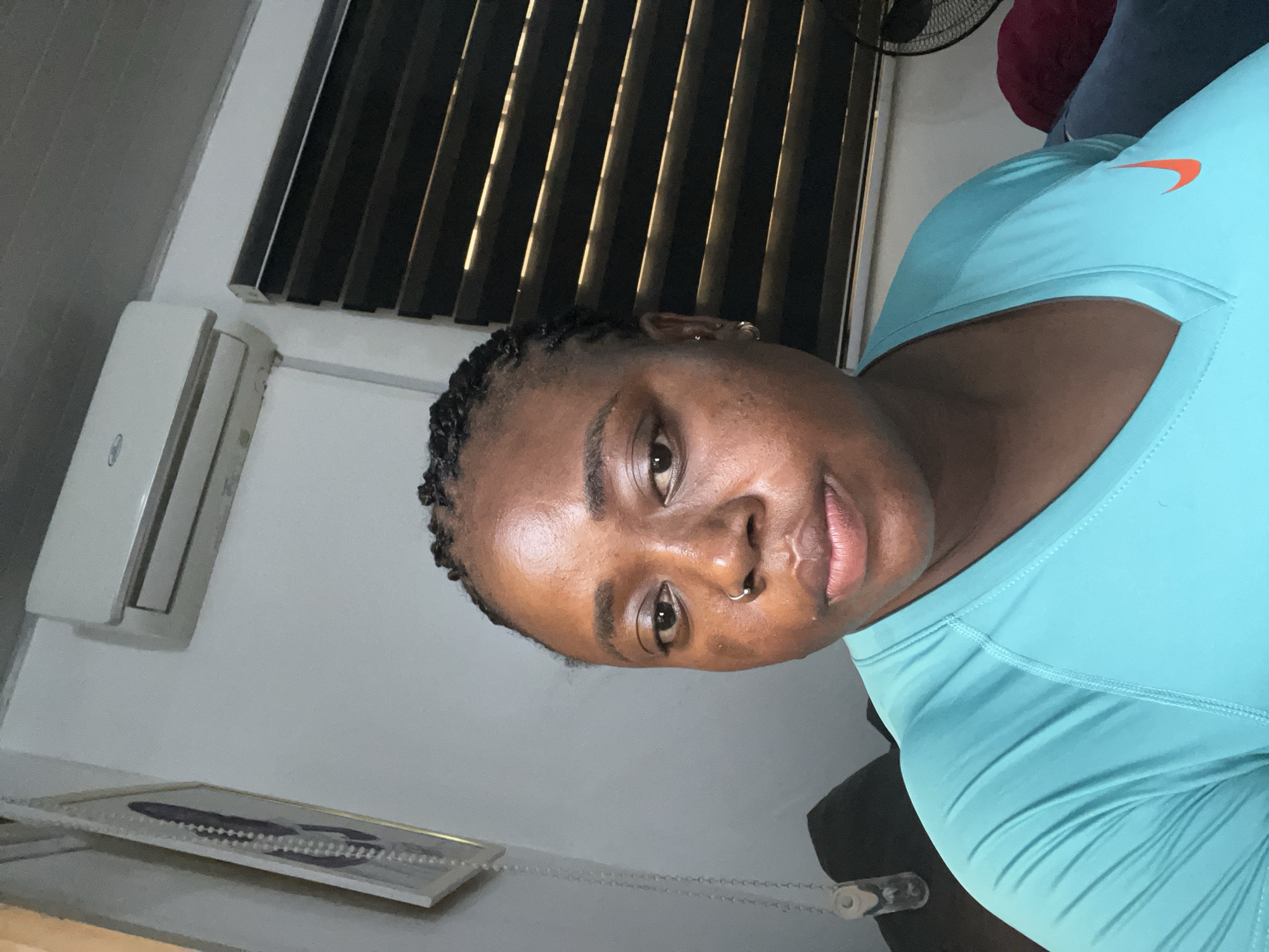 A dark-skinned woman with braided hair and a nose ring is pictured in a close-up, indoor selfie. She is wearing a turquoise v-neck t-shirt with an orange Nike logo. The background suggests a residential interior, with a wall-mounted air conditioning unit visible above her head, and blinds covering a window to her right. A framed picture with decorative beads hangs on the wall to her left. The scene is likely in Abuja, Nigeria, given the presence of an air conditioner, common in the climate.