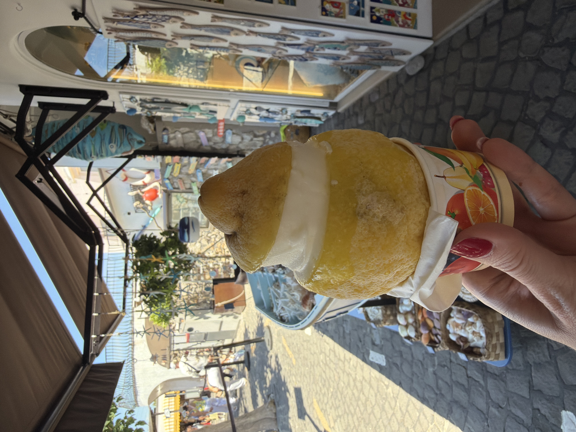 A first-person perspective shot depicts a person holding a partially eaten lemon granita, a frozen dessert, while standing in Anacapri, Italy. The granita, encased in a cup adorned with fruit illustrations, is held against the backdrop of a narrow street lined with shops and establishments. The street is paved with grey cobblestones and appears to be in a commercial area with pedestrians visible in the distance. The sky is bright, suggesting a sunny day. Buildings along the street feature open-air structures with awnings. One shop displays marine-themed merchandise, including blue fish decorations.