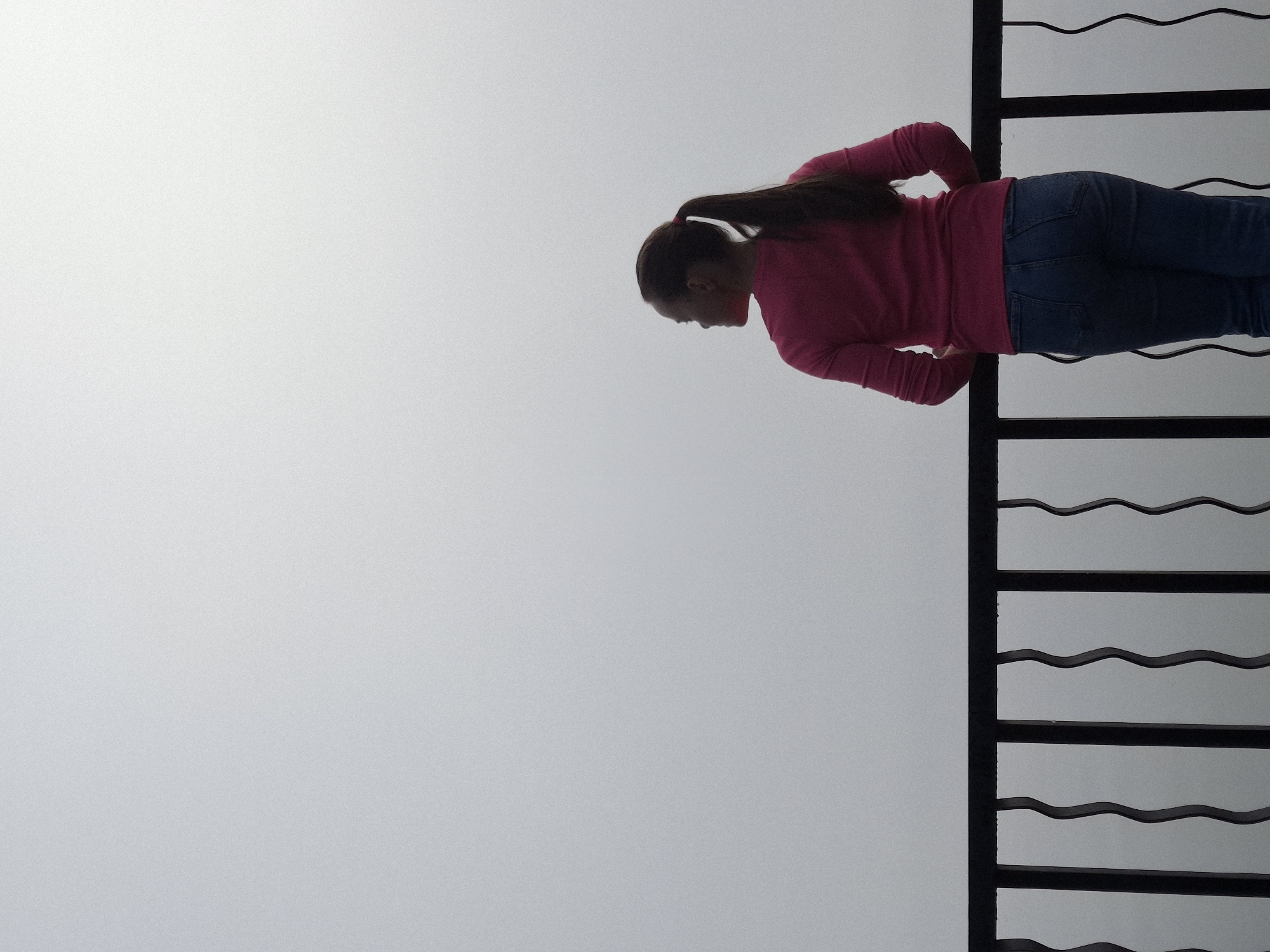 A woman with dark hair pulled into a ponytail stands with her back to the camera, leaning on a black metal railing. She is wearing a long-sleeved pink shirt and blue jeans. The background is a solid, overcast sky with no discernible features, suggesting a foggy or cloudy day. The railing is ornate, with vertical bars that have a wavy pattern. The image appears to be taken from an elevated position, possibly a balcony or viewpoint, overlooking a vast expanse of the sky. The location is not specified, but the context provided suggests it is in Chaikovsky, Russia.