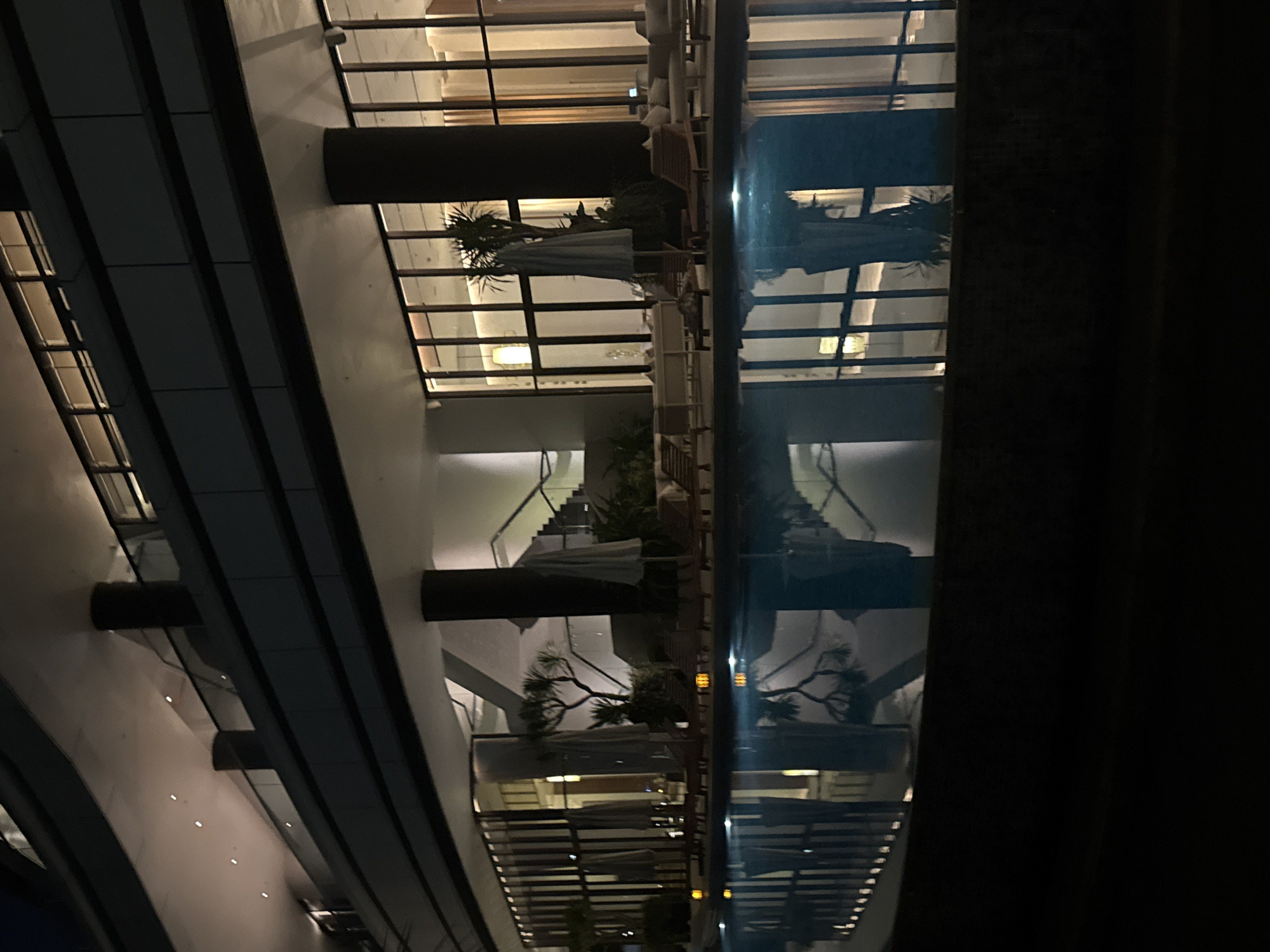 The image depicts a tranquil outdoor swimming pool area at night, likely part of a resort or upscale hotel in Bac Nha Trang Ward, Vietnam. The pool water is dark and reflects the ambient lighting from the building. Loungers and umbrellas are neatly arranged around the pool, suggesting a place for relaxation. The building itself features modern architecture with multiple levels, extensive glass windows, and prominent structural columns. Interior lighting spills out through the windows, illuminating the spaces within. The overall atmosphere is calm and sophisticated, characteristic of a high-end hospitality establishment.