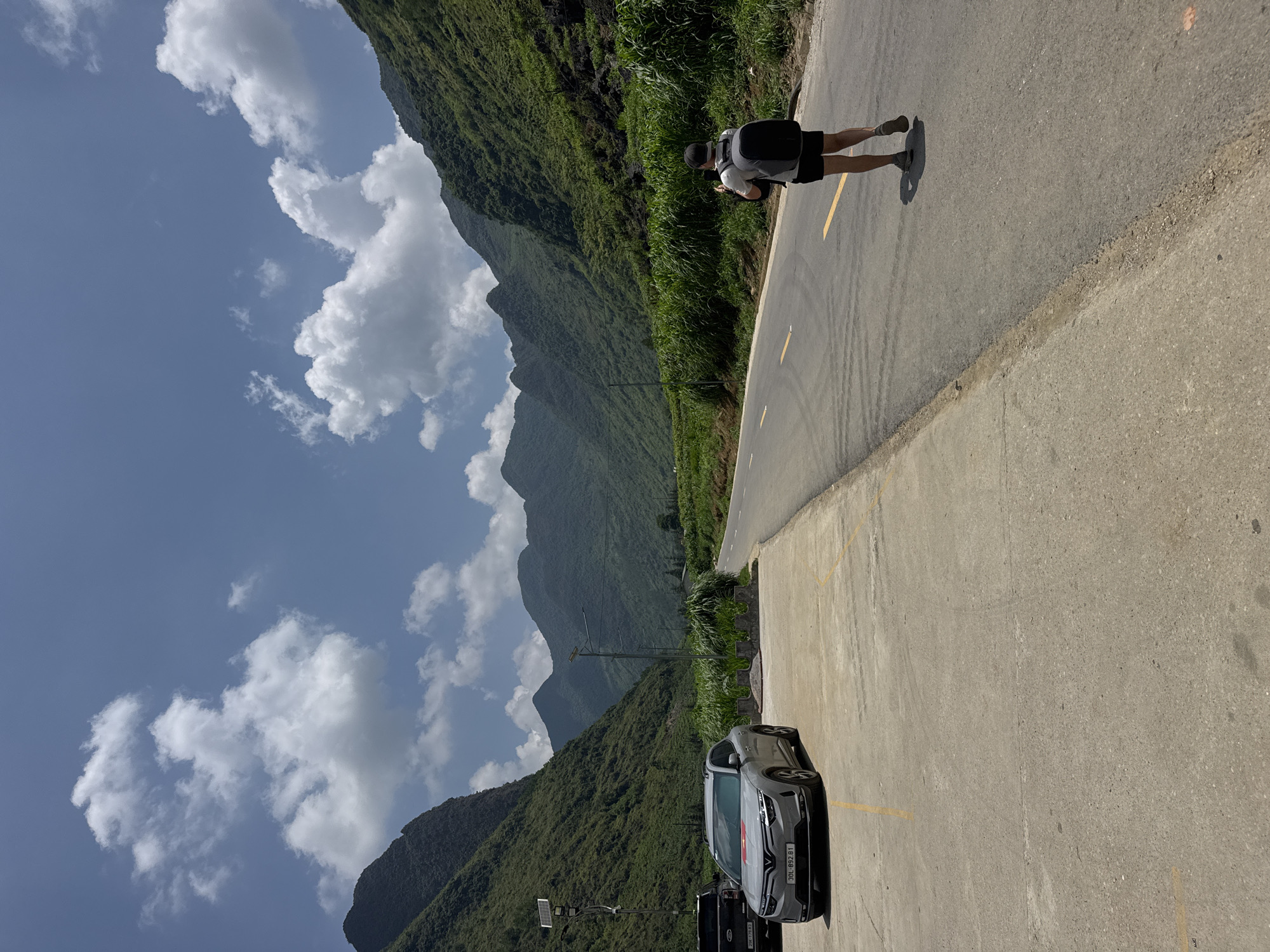 The image depicts a roadside scene with a striking mountainous backdrop in Mèo Vạc Commune, Vietnam. A man wearing a light-colored shirt, dark shorts, and a backpack stands on the road's edge, appearing to interact with a device or object he is holding. A gray car is parked on the roadside, positioned close to a street light. The road itself is asphalt, with a yellow line marking the edge. The mountains are lush with greenery and partially obscured by scattered white clouds in a bright blue sky. Power lines run along the road. The photo has a slight tilt, potentially due to the angle at which it was taken.