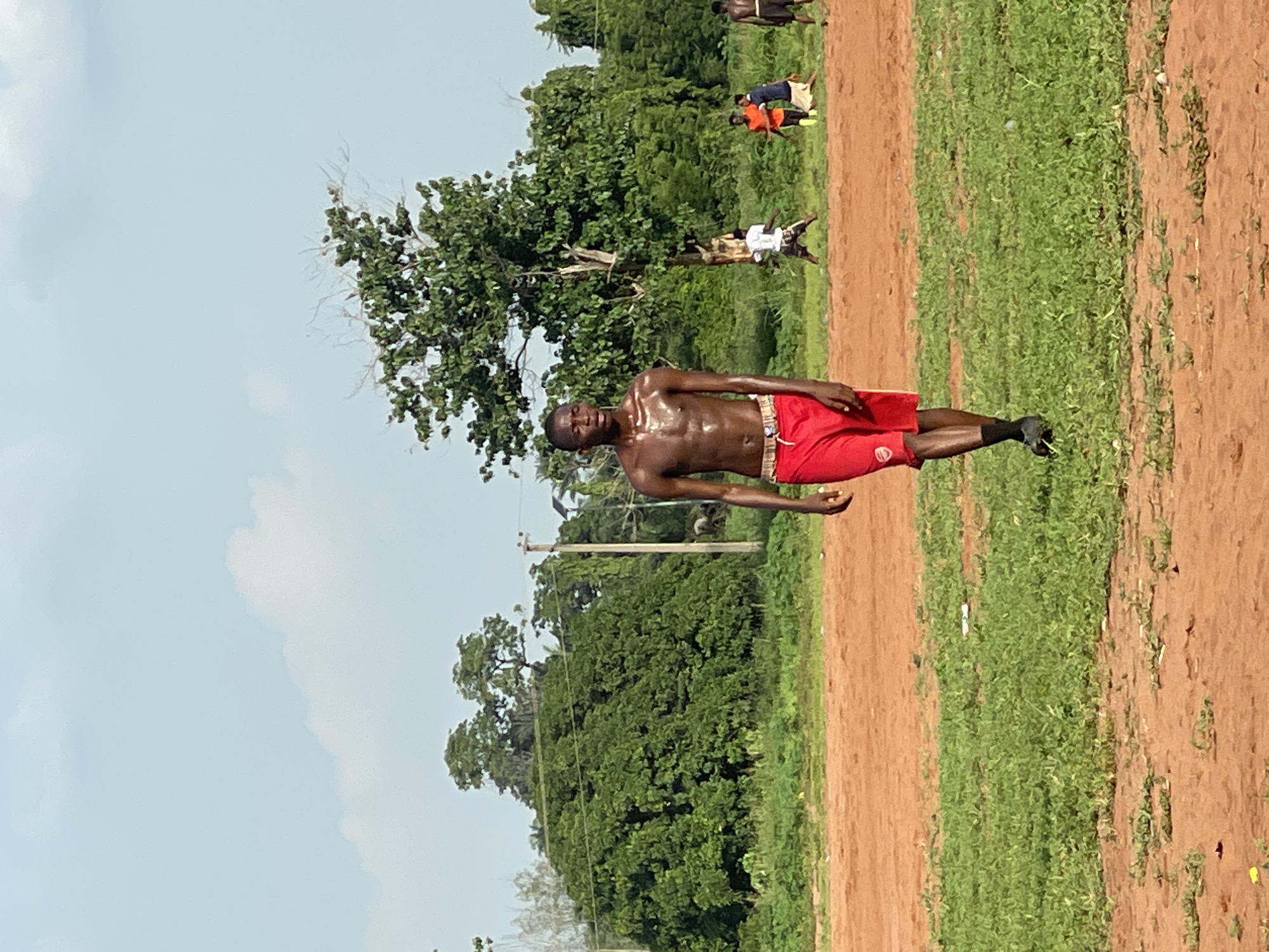 A shirtless man with dark skin, visibly sweating, stands upright in the foreground of an outdoor scene. He wears red athletic shorts with a small, indistinct white logo on the left leg, and dark socks and shoes. His physique appears muscular, indicating recent physical exertion. He faces slightly towards the right of the frame, looking towards the camera with a neutral expression.
The setting is an open field, characterized by stretches of reddish-brown earth alternating with patches of green, low-lying vegetation. In the mid-ground and background, several other individuals are scattered across the field, some standing, others possibly engaged in activity or resting. They are dressed in casual athletic wear, including t-shirts and shorts. Behind the field, a dense line of lush green trees forms a natural boundary, suggesting a wooded area or forest. Utility poles are visible among the trees. The sky above is bright and light blue with scattered white clouds, indicating clear, sunny weather, likely mid-day or early afternoon. The sweat on the main subject further suggests warm or hot conditions. The overall scene implies an outdoor sports or exercise event in a rural or semi-urban environment, consistent with the provided location context of Ogbogbo, Nigeria.