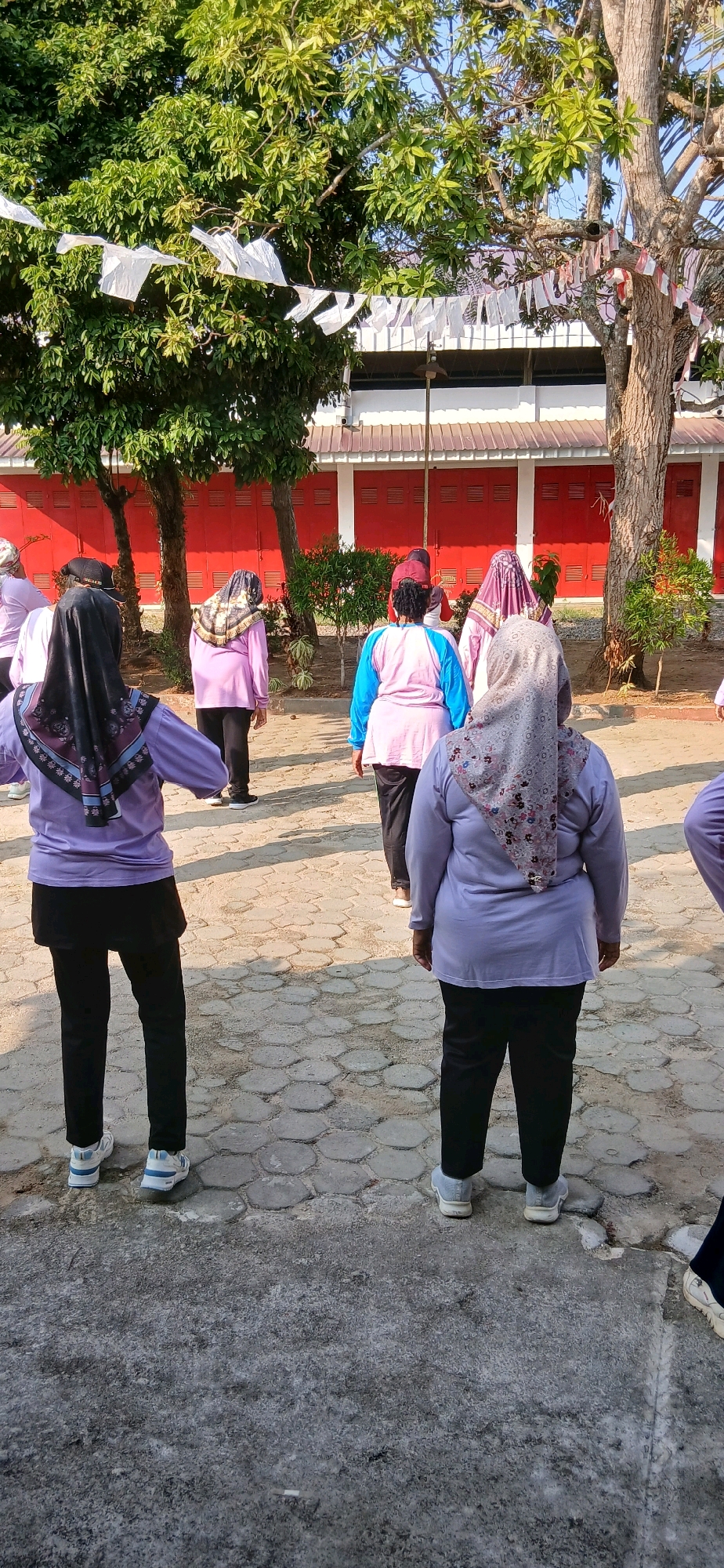 In Metro, Indonesia, a group of women are engaged in an outdoor exercise routine. They are positioned in a line facing away from the camera, suggesting a coordinated activity like aerobics or a morning walk. The women are dressed in modest athletic wear, primarily in shades of purple and pink, with some wearing hijabs. The ground is paved with interlocking hexagonal stones. In the background, a bright red building with white trim and a corrugated metal roof is visible, flanked by trees with lush green foliage. White pennant-like flags are strung across a wire between trees. The scene appears to be a public space, possibly a schoolyard or a community park, bathed in the bright sunlight characteristic of the region.