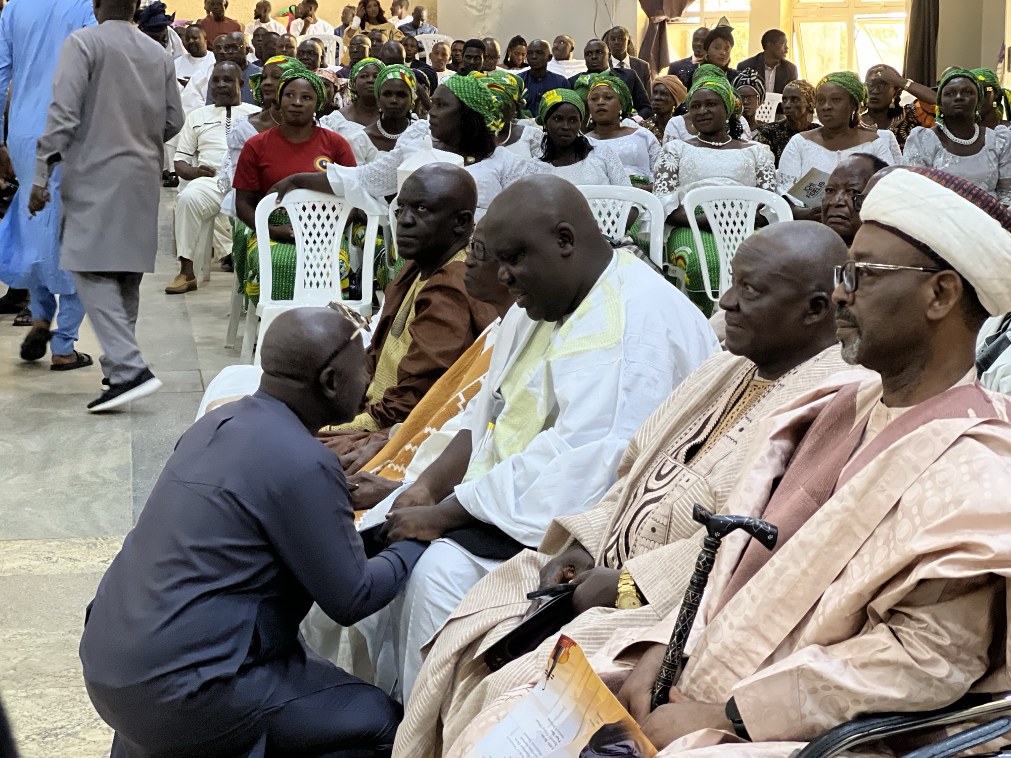 The image shows a gathering of people in what appears to be an indoor event, possibly a ceremony or religious service, in Kabong, Nigeria. The foreground is dominated by several men, some in traditional attire. To the left, a man in a dark blue kaftan is kneeling. In the center, a man in a white kaftan with intricate embroidery is seated, looking down. To his right, another man in a similar light-colored traditional outfit is also seated, with a gold watch visible on his wrist. A decorative cane rests in front of him.
Behind these men, a crowd of people, predominantly women, are seated on white chairs. Many of the women are wearing vibrant headscarves and traditional clothing in white and green. Some men are also visible in the background, seated and observing. The lighting suggests an indoor environment, possibly with natural light coming from windows that are not fully visible in the frame. The overall atmosphere appears formal and respectful.