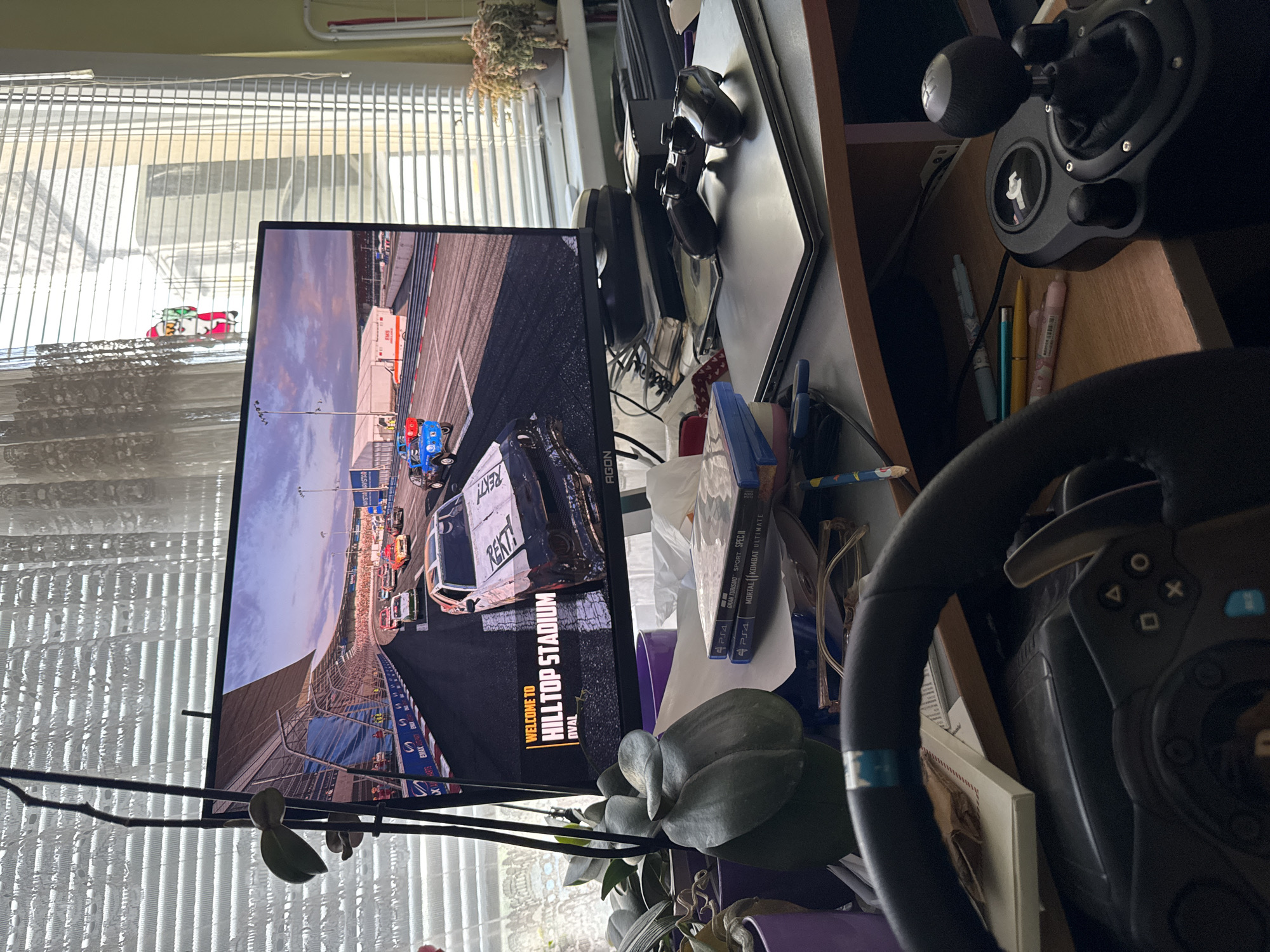 The image depicts a gaming setup in a room with white Venetian blinds. The blinds are slightly open, allowing some daylight to illuminate the room. On a desk, there's a computer monitor displaying a racing game. The game scene is set at "HILLTOP STADIUM", as indicated on the screen. A white racing car with the word "REKT" on it is prominently visible on the monitor.
To the right of the monitor, there is a black gaming steering wheel and a gear shift. A PlayStation 4 (PS4) game case is visible. Other items on the desk include pens, glasses, and an unidentified plant.