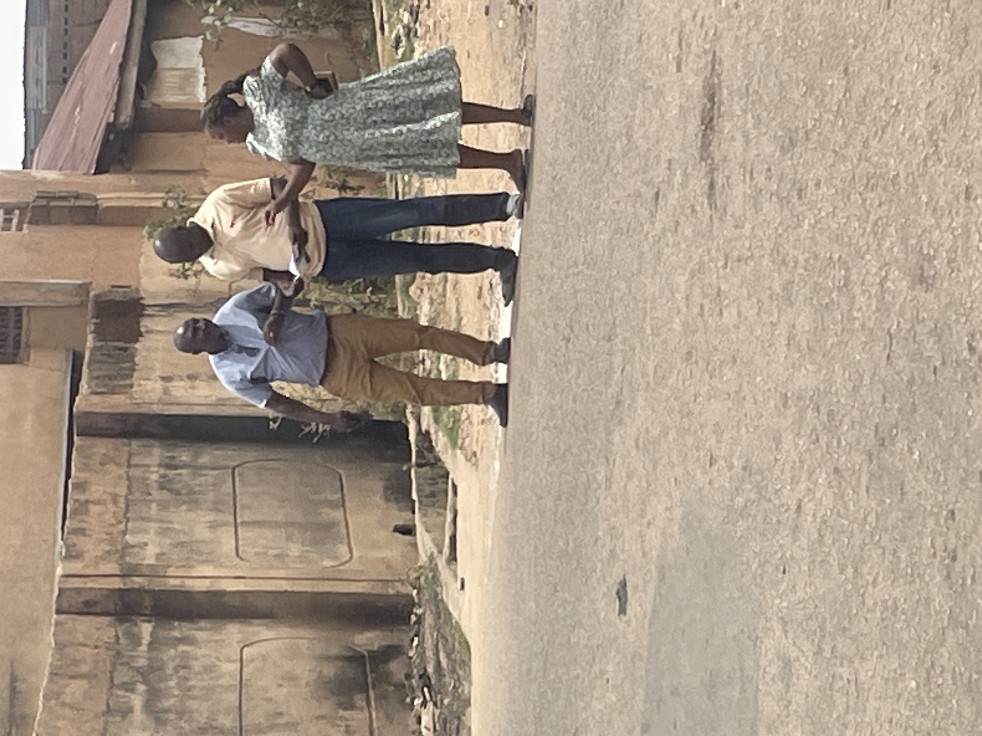 Three people are standing on a street in a residential area of Jos, Nigeria. The person closest to the camera is wearing a light blue shirt and khaki pants. The person in the middle is wearing a tan shirt and jeans. The person furthest from the camera is wearing a patterned dress. The street is paved and there is a building in the background.
