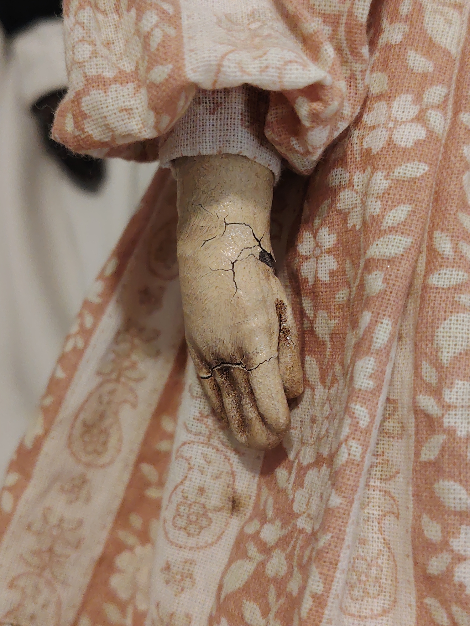 A close-up vertical shot focuses on the weathered hand and patterned clothing of what appears to be an antique doll. The doll's right hand, made of a light, possibly wooden or composite material, shows significant aging, characterized by numerous fine and deep cracks across its surface, especially prominent around the wrist and palm. Some of these cracks are filled with darker material, suggesting wear or dirt accumulation over time. The fingers are delicately sculpted, and there's a visible roughness or texture to the material.
The hand emerges from a light-colored, ruffled sleeve cuff that is adorned with a repeating peach or light orange floral or leaf pattern. Beneath this outer sleeve is a plainer, tighter-fitting inner cuff of off-white fabric. The main body of the doll's attire, likely a skirt or dress, features a different pattern. It consists of alternating vertical stripes: one stripe is solid light cream or off-white, while the adjacent stripe has a peach or light orange repeating paisley or intricate floral design. Scattered floral or leaf motifs also appear on the lighter stripes. The fabric of the clothing appears to be a woven material, possibly linen or cotton, with visible texture.
The background is out of focus, suggesting a shallow depth of field, and appears to be a plain, light surface, likely indoors. The overall lighting is soft and even, highlighting the textures and details of the doll and its attire. No specific activity is depicted, and there is no visible text.