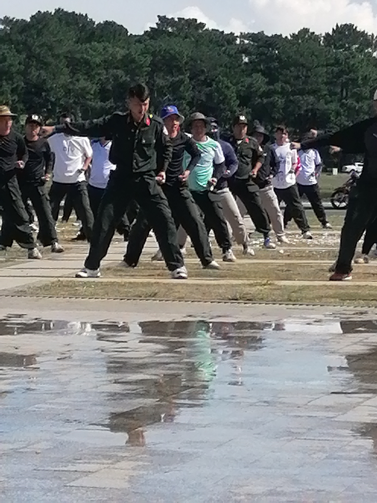 A group of people, some in military-style uniforms, are performing a synchronized exercise outdoors. The individuals are spread across a paved area, with trees and a cloudy sky visible in the background. The foreground shows a wet, reflective surface, possibly puddles on the pavement. The people appear to be in motion, with arms extended and legs in various positions, suggesting they are engaged in a coordinated physical activity. Some individuals are wearing hats, including a blue baseball cap and a brimmed hat. The lighting indicates it is daytime. There is no visible indication of the city of Da Lat, Vietnam.