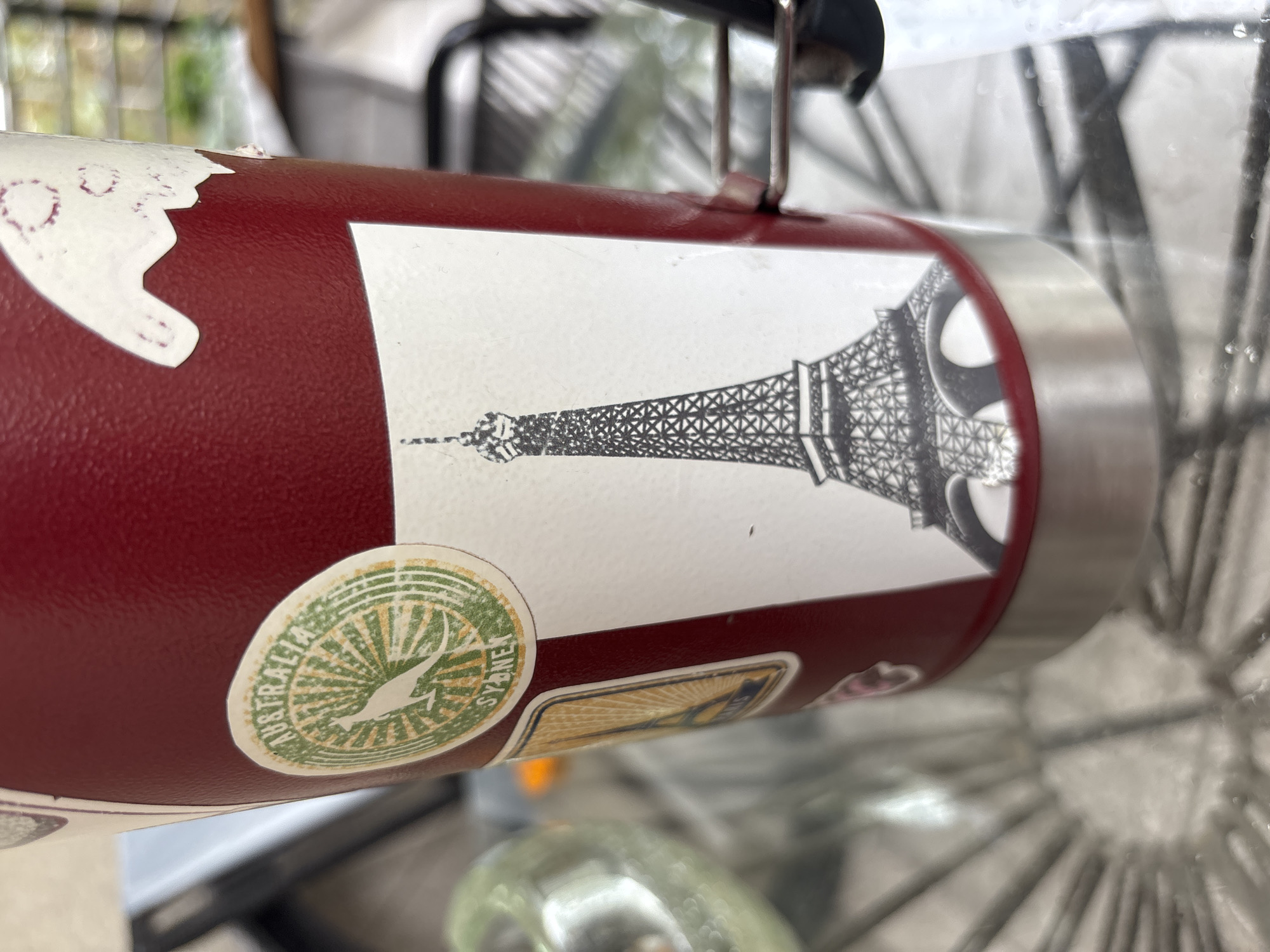 A maroon water bottle with several stickers is placed in front of a blurred background that seems to be a bicycle wheel. The bottle has a white sticker with a black and white illustration of the Eiffel Tower. There is also a round green sticker with a kangaroo silhouette and the word "Australia" and "Sydney". There's also another partially visible rectangular sticker below the kangaroo sticker. The location in the city of Buenos Aires, Argentina, cannot be determined from this image.
