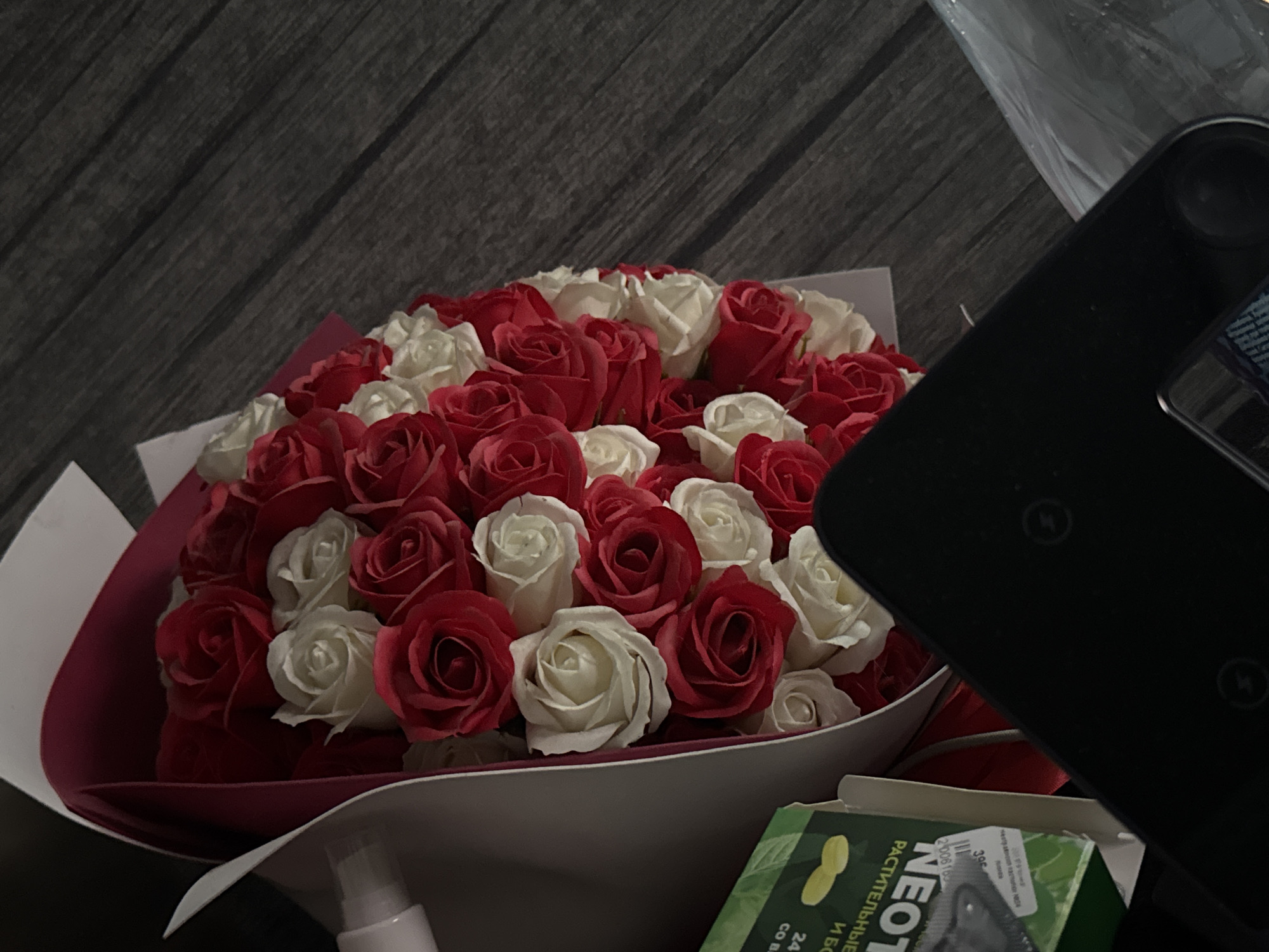 The image shows a bouquet of red and white roses arranged in a bouquet with a red and white wrapping. The bouquet is placed on a dark wooden surface, possibly a floor or table. To the left of the bouquet, a green box with some text and images is visible. A black object, possibly an electronic device, is in the foreground, obscuring part of the bouquet and the wooden surface. To the right, a translucent plastic bag is present. The lighting appears to be dim, with some reflections on the black object. The overall scene suggests a domestic or retail setting. There are no people visible in the image.