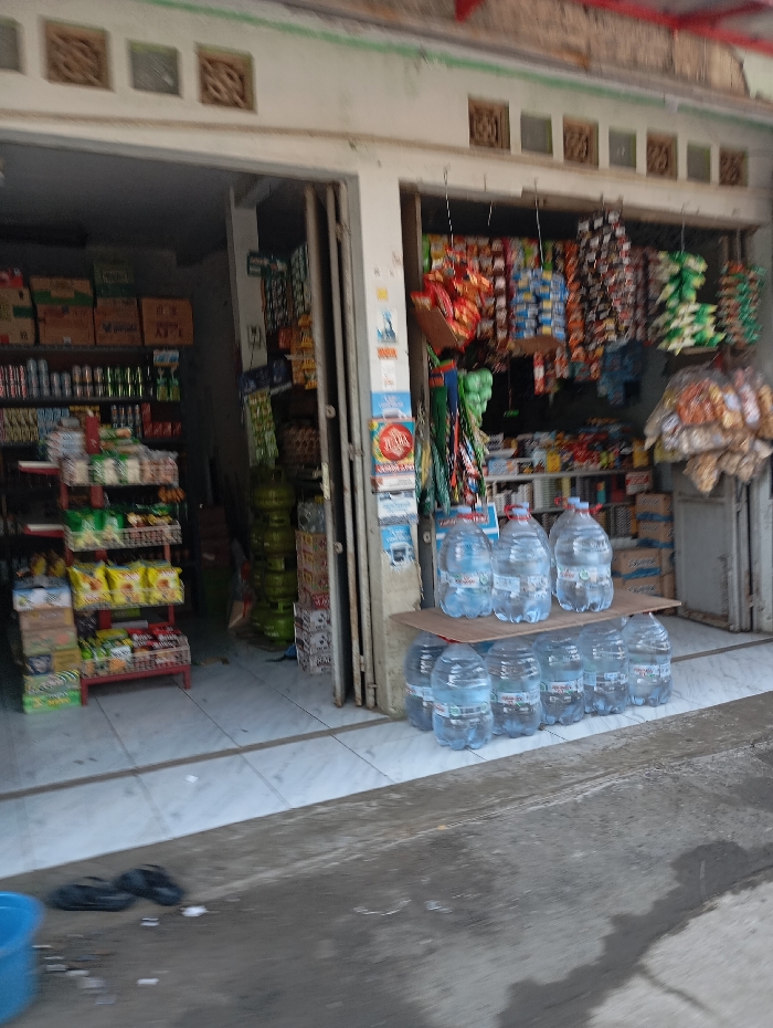 The image displays the storefront of a small market or convenience store located in Cipatik, Indonesia. The building features two open entrances, revealing white tiled flooring inside. Above the doorways, decorative square vents are visible on the building facade.
Outside the right entrance, a two-tiered wooden stand holds eleven large, clear plastic bottles of water; five bottles are on the upper tier and six are on the lower. Numerous small, individually packaged goods, contained in colorful plastic bags, hang suspended from lines across the upper portion of this entrance.
Inside the left entrance, shelves are stocked with various packaged products and beverages. Multiple green LPG gas cylinders are visible stacked deeper within the store. Inside the right entrance, additional shelves are stocked with assorted merchandise.
On the tiled ground directly in front of the left entrance, a pair of black sandals are present. A light blue plastic bucket is partially visible in the lower left corner of the frame. The immediate foreground consists of a darker, rough paved surface, exhibiting scattered wet patches. No individuals are visible in the scene.