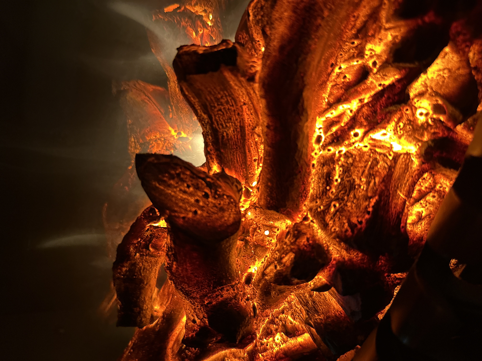 This is a close-up, dimly lit, macro photograph of burning logs in a fireplace. The logs are a deep orange and red, with some appearing to be glowing embers. There is smoke or steam rising from the burning material, creating a hazy atmosphere. The background is dark and out of focus. The photograph appears to be taken from a low angle, looking up into the fire.