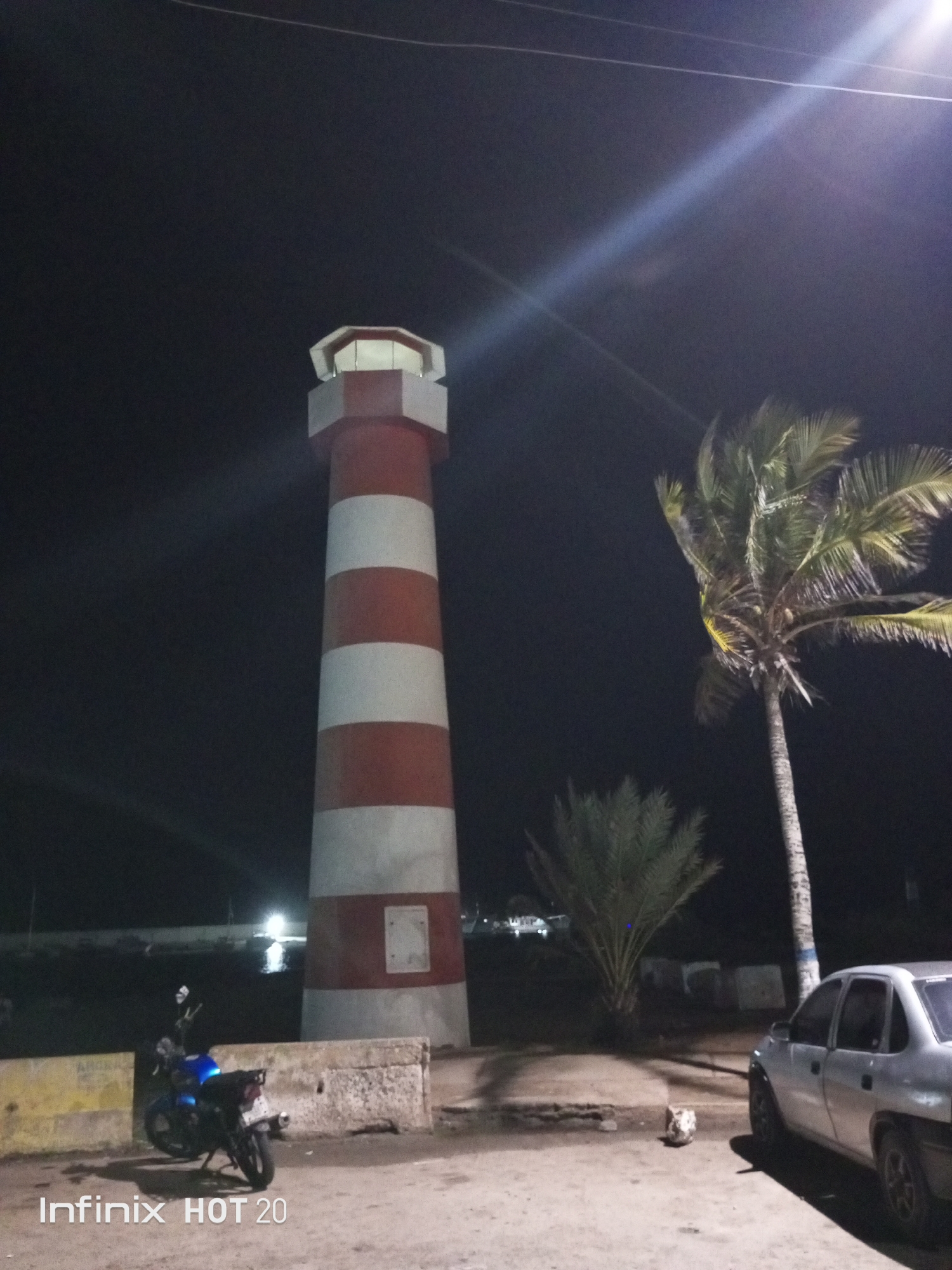 The image captures a nighttime scene in what appears to be a coastal or port area, specifically Porlamar, Venezuela, given the context. The dominant feature is a tall lighthouse, painted with alternating red and white horizontal stripes, standing prominently slightly to the left of the center. Its top section is illuminated, suggesting it's operational.
To the right of the lighthouse, a tall palm tree with long, green fronds extends upwards, its foliage partially catching the light from an unseen source, creating a slight glow. Beneath it, another, shorter palm tree is visible.
In the foreground, a blue motorcycle is parked on the left side, behind a concrete barrier or wall. On the right, the rear and side of a silver car are visible, parked on what looks like a dirt or uneven paved surface. Near the car, a small, light-colored object rests on the ground.
The sky is completely dark, indicating it is night. Several bright lens flares are visible in the upper right portion of the image, suggesting a strong artificial light source just out of frame or directly facing the camera. Distant lights can be seen across a body of water in the background, to the left of the lighthouse. The scene is still and quiet, without any visible people or activity.
A watermark "Infinix HOT 20" is present in the bottom left corner of the image.