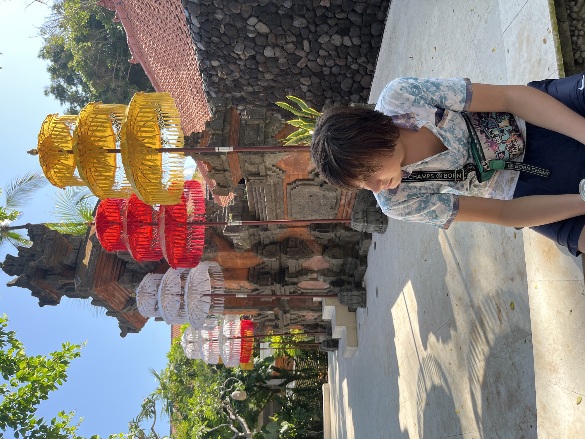 In Kuta Selatan, Indonesia, a young person with brown hair sits outdoors on a light-colored tiled surface. They are wearing a light-colored, short-sleeved shirt with a blue and white floral pattern and dark blue shorts. A green fanny pack with a colorful graphic design is worn across their chest. To the left of the person are several tiered ceremonial umbrellas. The umbrellas are arranged in rows and vary in color, including white, red, and yellow. Behind the umbrellas, there is an ornate stone structure, possibly a temple or traditional building, with carved details and a tiled roof. The background features lush green trees and a clear blue sky. The ground is illuminated by sunlight, casting shadows from the umbrellas and foliage.