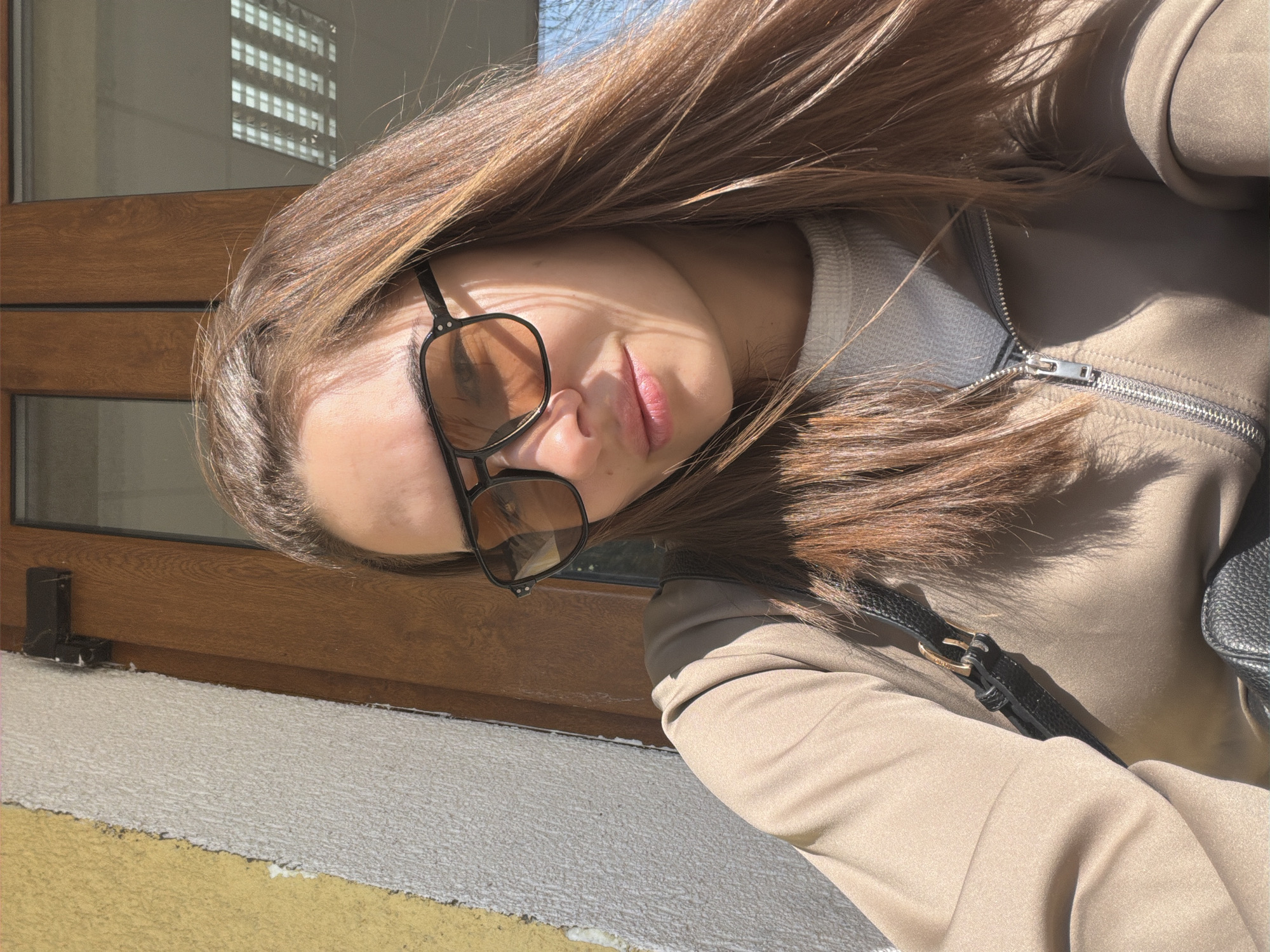 A young woman is posing for a selfie in Sofia, Bulgaria. She is wearing a beige zip-up jacket over a light-colored top, with a black handbag strap visible on her shoulder. Her long, brown hair falls around her face, and she is wearing square, tinted glasses. Sunlight is casting shadows across her face and clothing. The background includes a window with a distinctive grid pattern, possibly reflecting another building. A wooden frame surrounds the window. The exterior area below the window appears to be a wall with a textured surface.
