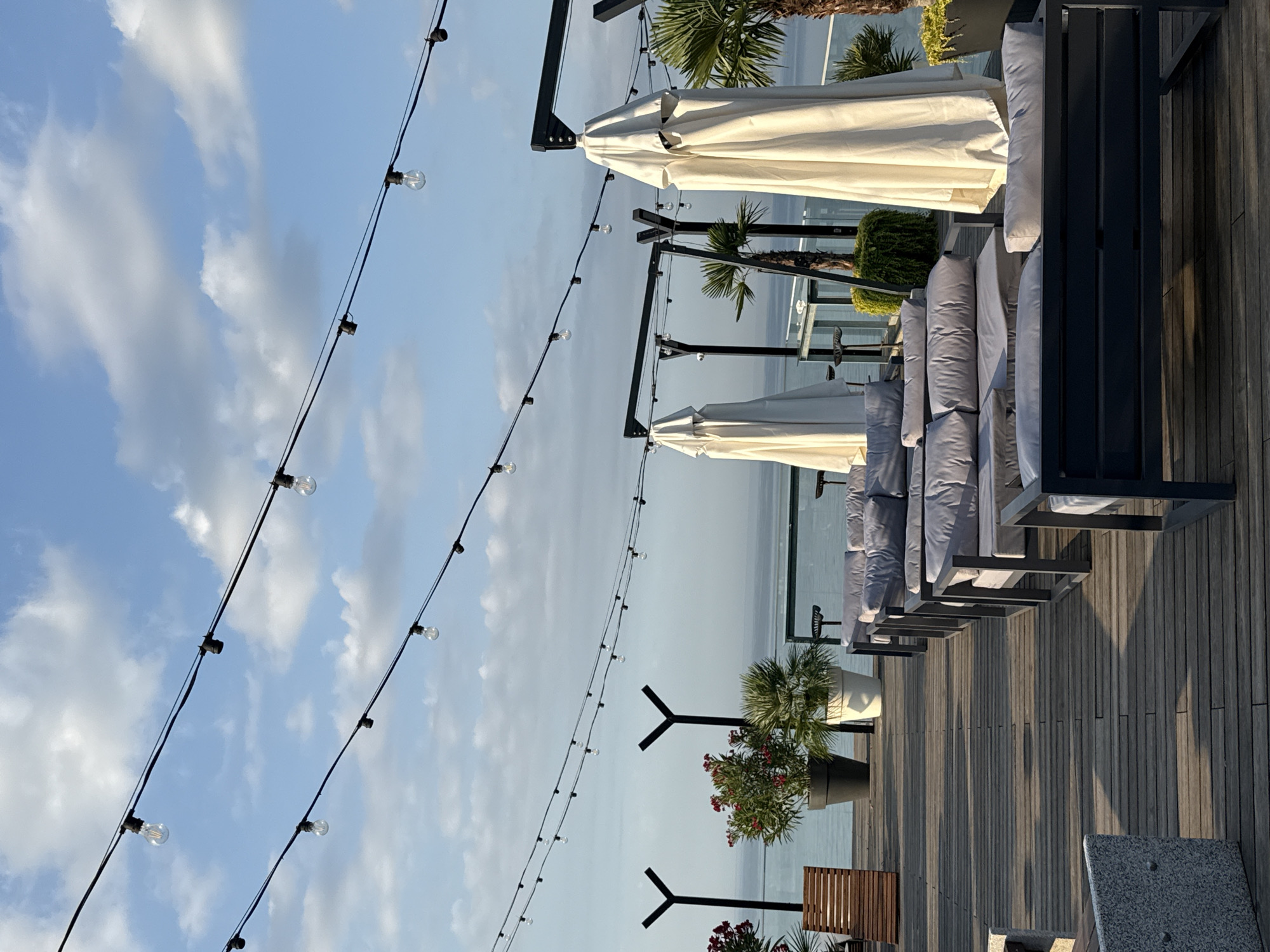 The image shows an outdoor seating area with lounge furniture and large potted plants on a wooden deck overlooking a body of water. String lights with exposed bulbs are suspended overhead. Two white, closed umbrellas stand prominently. The sky is blue with scattered white clouds. The scene suggests a restaurant or cafe with a view. The location is identified as Yalta, Ukraine.