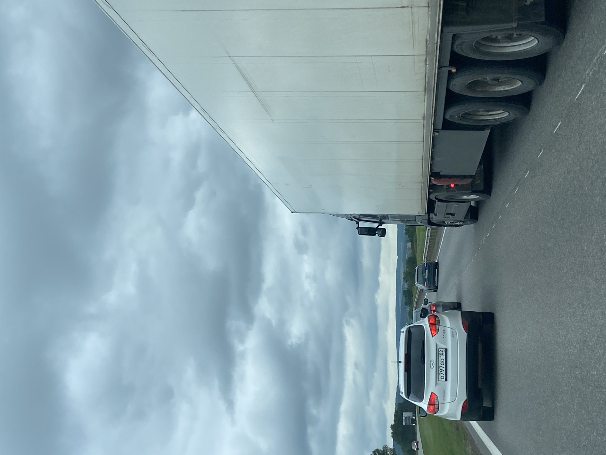 The image depicts a cloudy day on a highway in Uzytamak, Russia. The foreground shows the rear of a white Hyundai SUV with a license plate reading "0797co 102". To the right of the SUV, a large white semi-trailer truck occupies a significant portion of the frame, its trailer extending towards the right edge. In the middle ground, other vehicles, including a dark-colored sedan, are visible on the highway. The background consists of a tree line and rolling hills under a heavily overcast sky. The highway appears to have multiple lanes, indicated by dashed white lines.