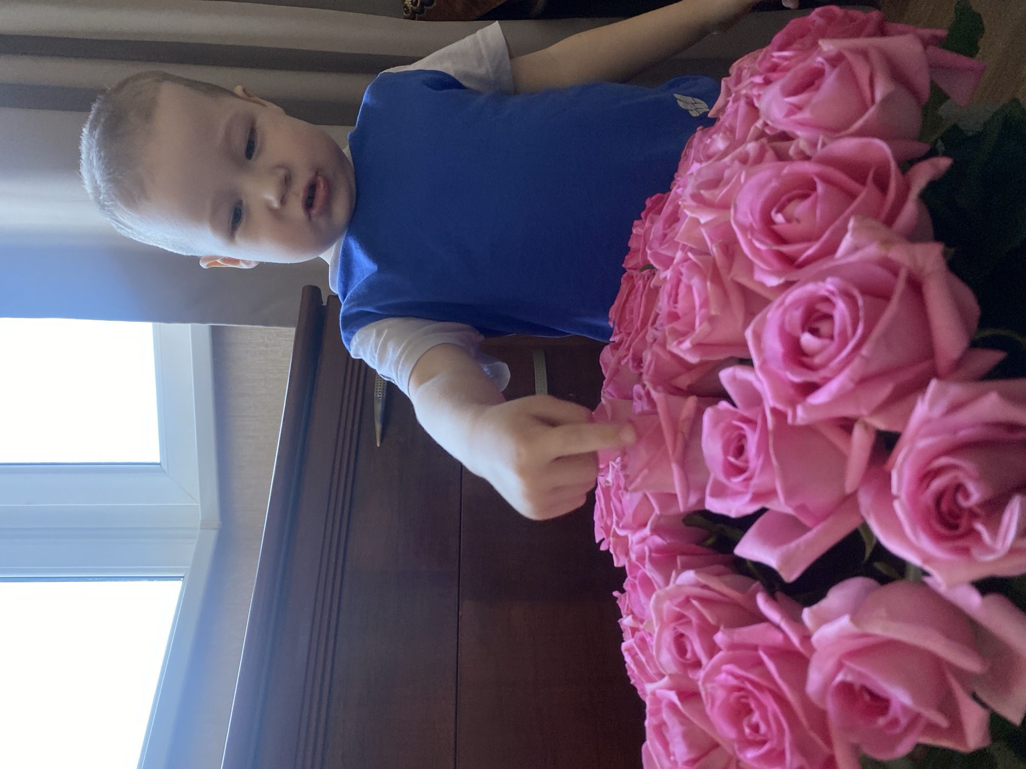 A young child in a blue and white shirt is reaching toward a large bouquet of pink roses. The roses are densely clustered together. The child is standing in front of a brown wooden dresser. A window with white frames is visible in the background. The location is Sochi, Russia.