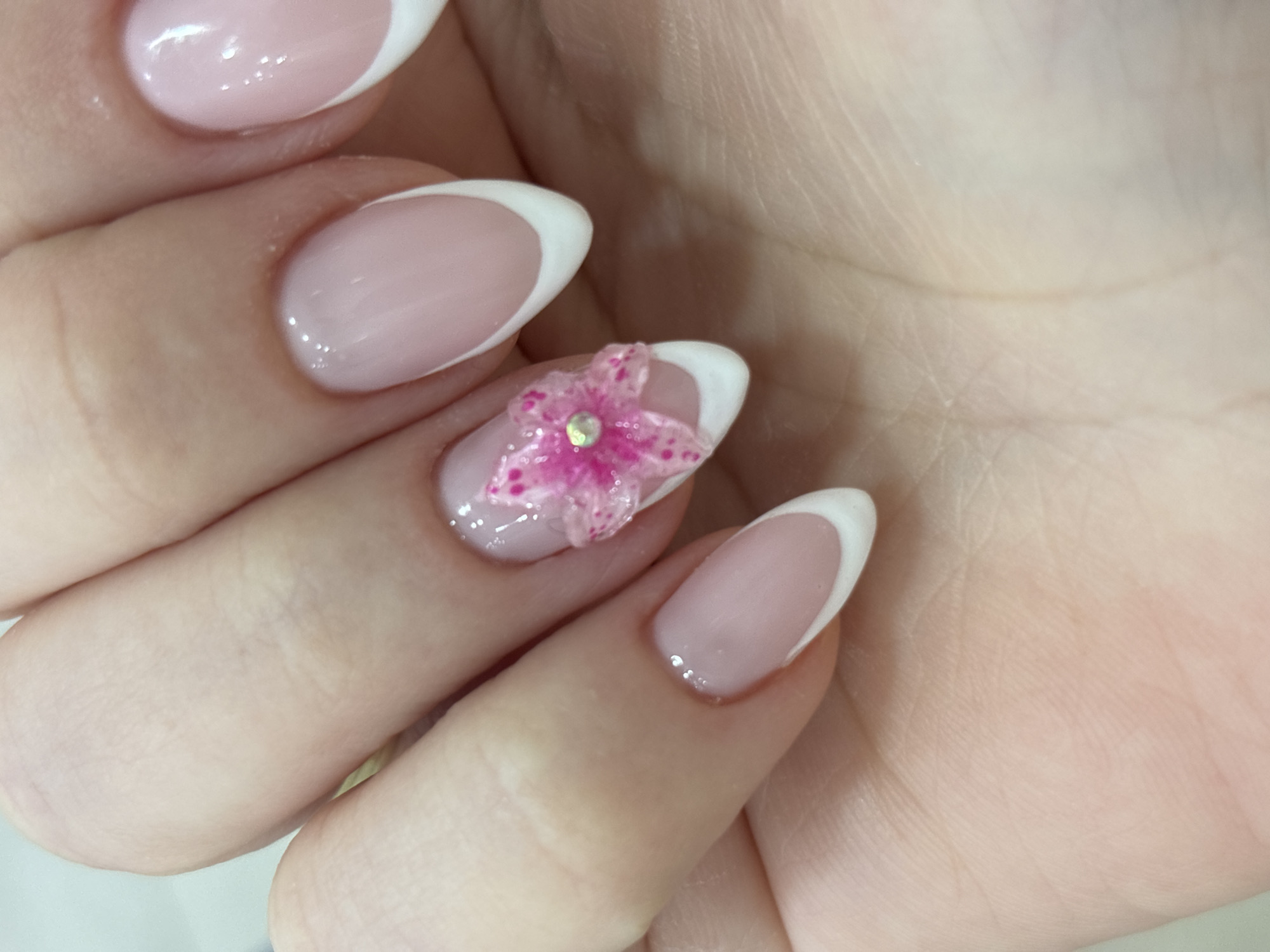 A close-up shot shows a hand with manicured nails. The nails have a French manicure design with a pale pink base and white tips. One nail is adorned with a pink flower with a rhinestone in the center. The hand is resting against another part of the hand. The background is blurred. The photograph was taken in Sofia, Bulgaria.