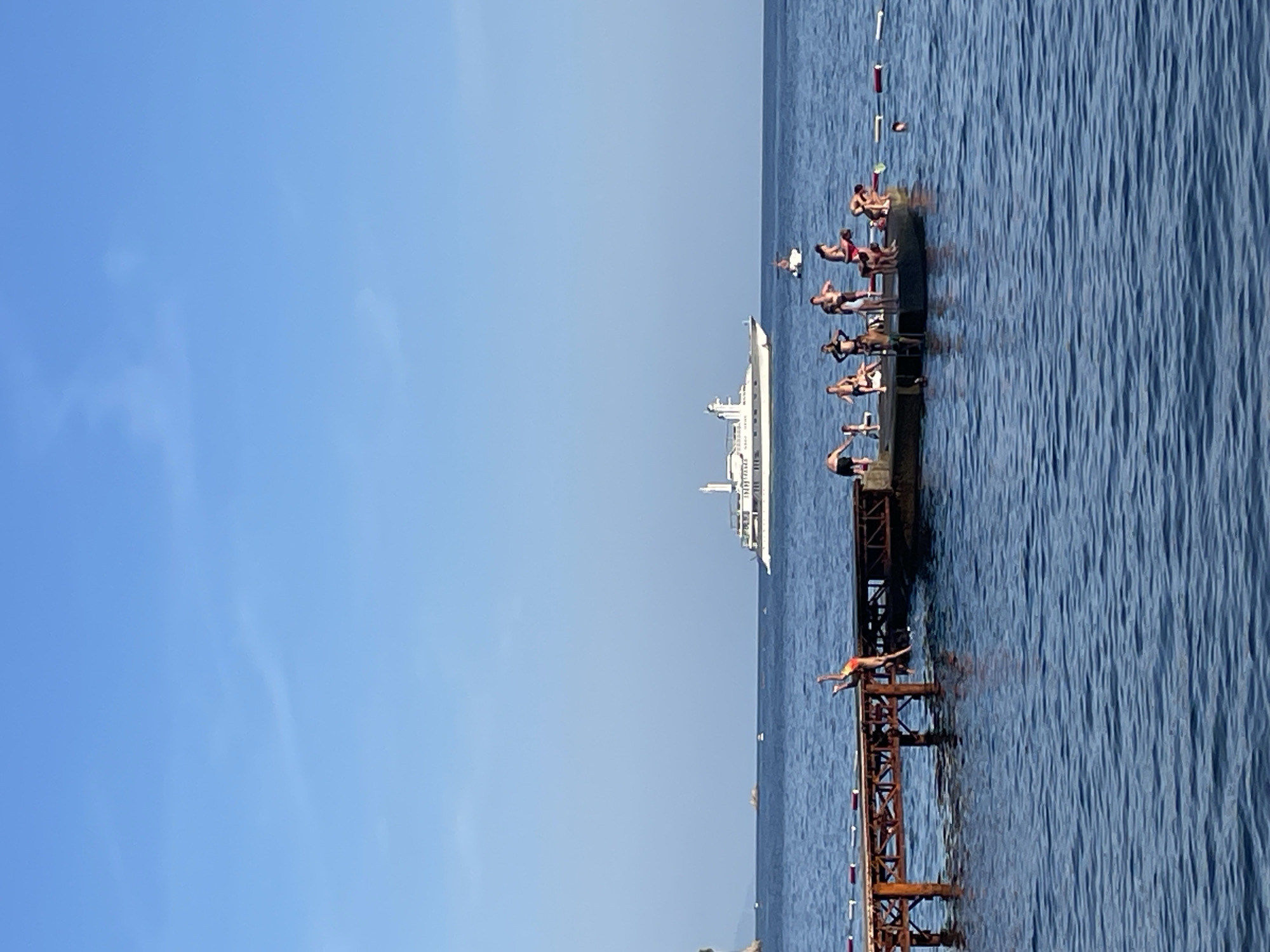 A wide shot captures a bright, clear day at sea, with a large white yacht sailing in the distance. In the foreground, a wooden pier juts out into the calm blue water. A group of people are gathered on the pier, with some standing and others in various poses, seemingly preparing to jump or dive into the sea. One individual is mid-air, leaping from the pier. Further out in the water, a few more people are visible, one on a small boat or flotation device and another swimming. The scene suggests a leisure activity on a sunny day.
