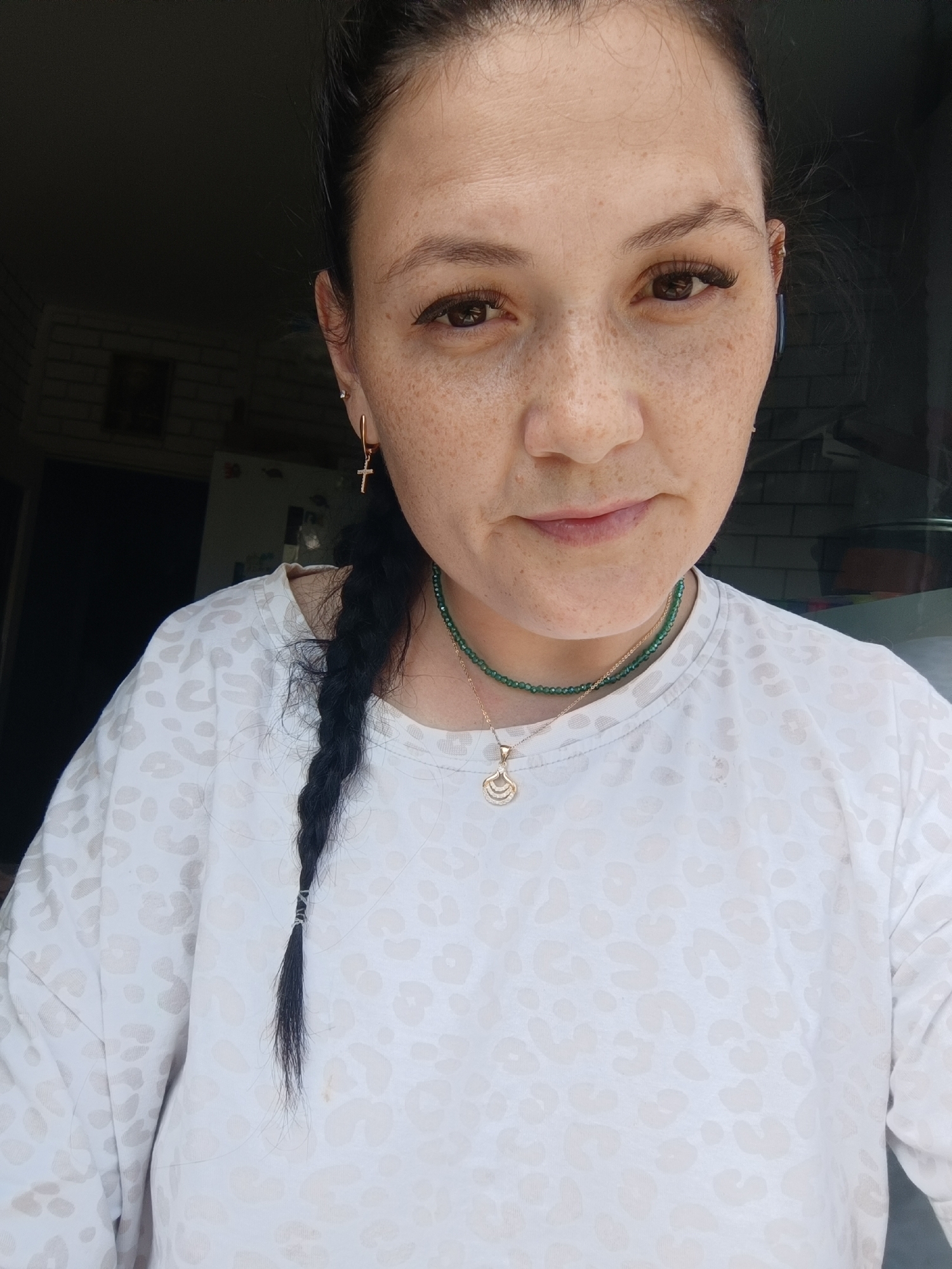 A medium shot shows a Caucasian woman with brown hair pulled back into a braid, dark brown eyes, and fair skin with freckles. She is wearing a white shirt with a subtle light gray leopard print pattern, a green beaded necklace, and a gold pendant. She also has a cross earring in her left ear. The background is dark with some brickwork visible on the left and a hint of a refrigerator on the far left. The woman is looking directly at the camera. The image appears to be taken indoors, possibly in a kitchen or hallway.
