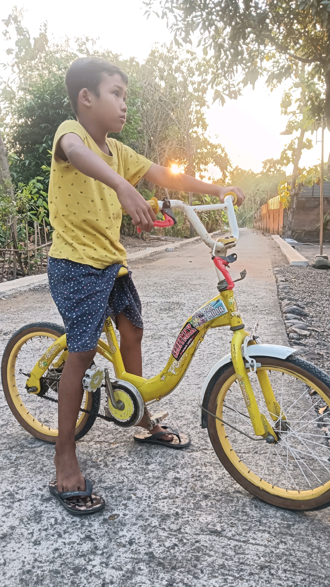 A young boy stands with his bicycle on a concrete path in Tanggan, Indonesia. The boy is wearing a yellow t-shirt and blue shorts. The bicycle is yellow and has white handlebars. The sun is setting in the background, casting a warm glow over the scene. The path is lined with trees and houses.
