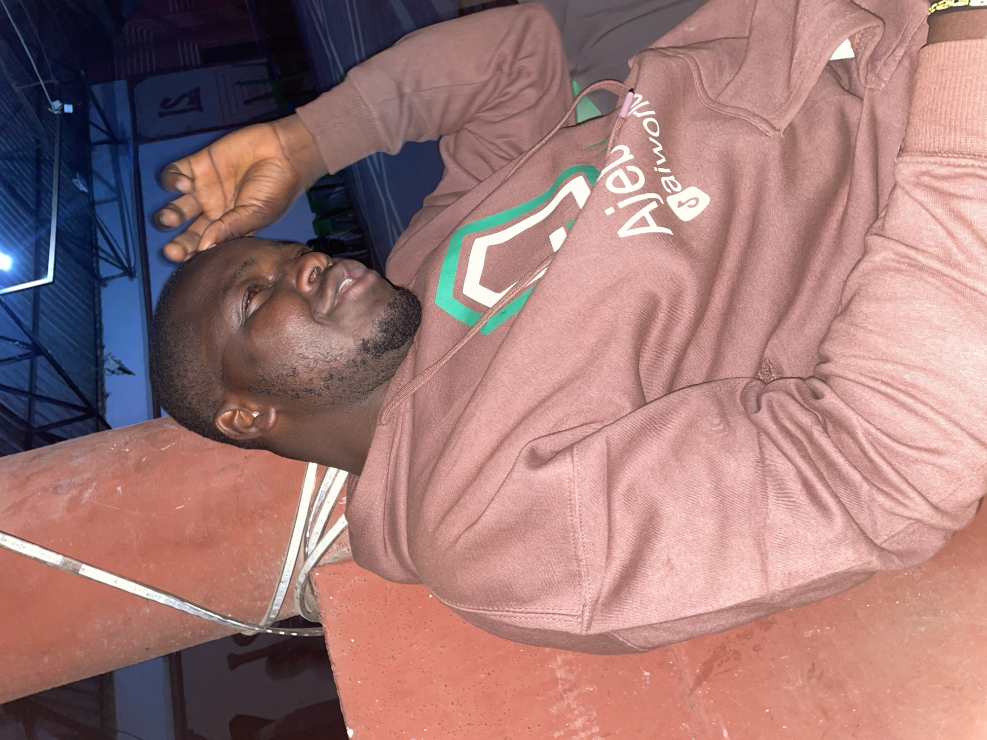 A man lies on a red tiled surface. He is wearing a brown hoodie. His hand is resting on his forehead. He is dark-skinned and has a beard. The background is blurry and appears to be nighttime. The location is potentially Jos, Nigeria.