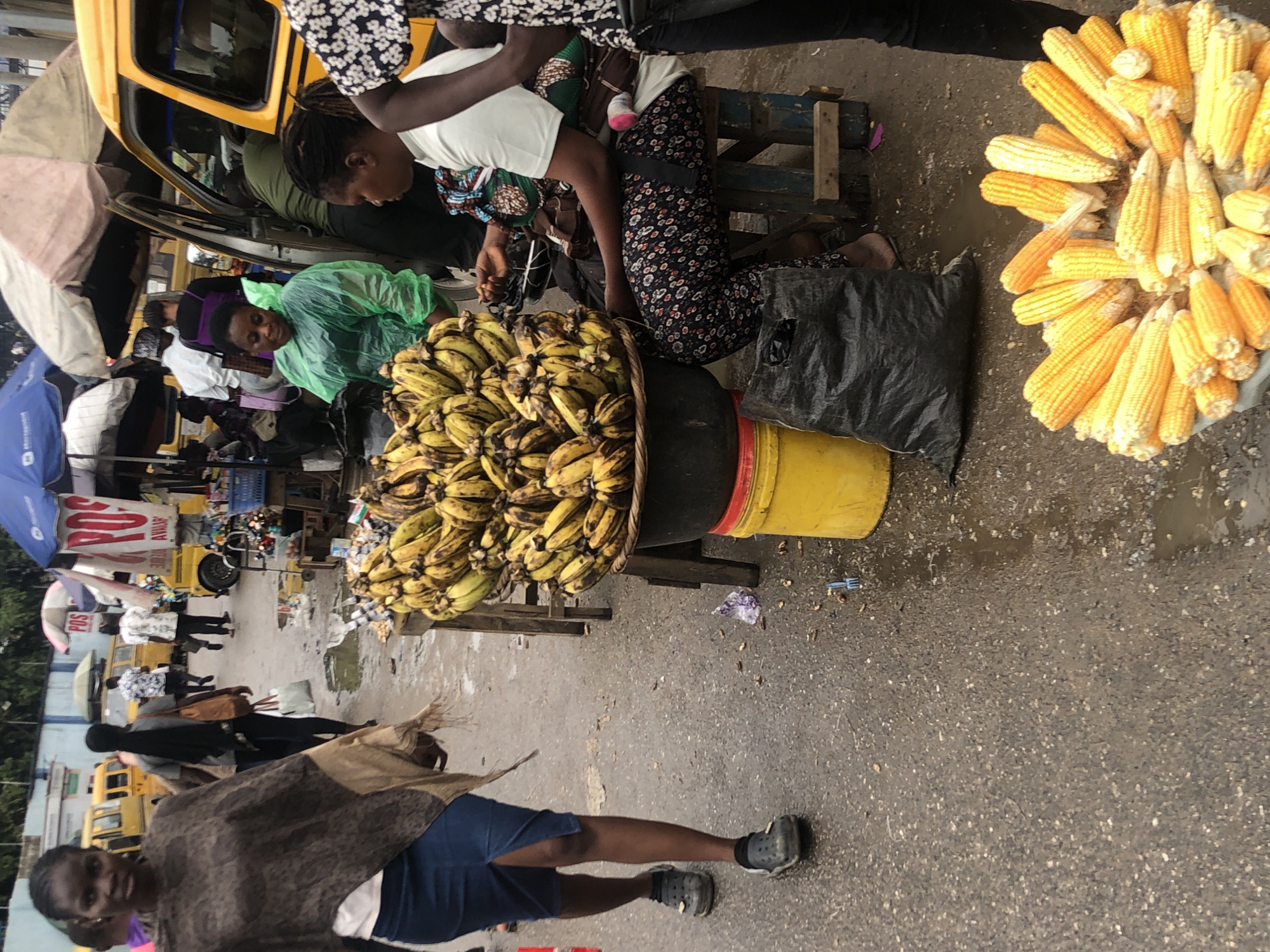 The image depicts a street scene in Ikeja, Nigeria, likely a market area. A woman walks towards the viewer on the left, wearing a patterned poncho and a blue skirt. In the background, several yellow minibuses are parked, a common mode of transportation in Lagos. To the right, a stall displays a large quantity of bananas in a black basin. Next to the banana stall, a woman is seated, appearing to be tending to a baby wrapped in a fabric sling. She wears a white t-shirt and a patterned skirt. Another woman, wearing a green plastic poncho, stands near the minibus. In the foreground, a tray is filled with corn on the cob, ready for sale. The ground is wet and appears to be a paved street with puddles, suggesting it may have recently rained or is a low-lying area. The overall atmosphere suggests a bustling urban marketplace.