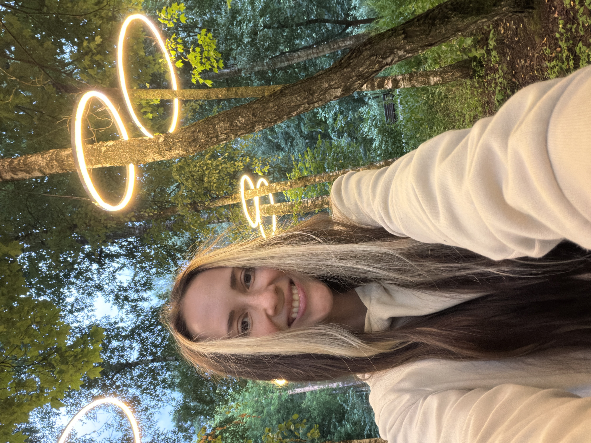 A young woman with long, dark brown and blonde highlighted hair is taking a selfie outdoors in a wooded area. She is smiling broadly, looking directly at the camera. She is wearing a cream-colored hooded sweatshirt. In the background, trees with white bark are visible, and several circular, illuminated lights are suspended from the branches, emitting a warm orange glow. The foliage is lush and green, suggesting it is a natural setting within the city of Malakhovka, Russia. The lighting suggests it is either dusk or twilight.